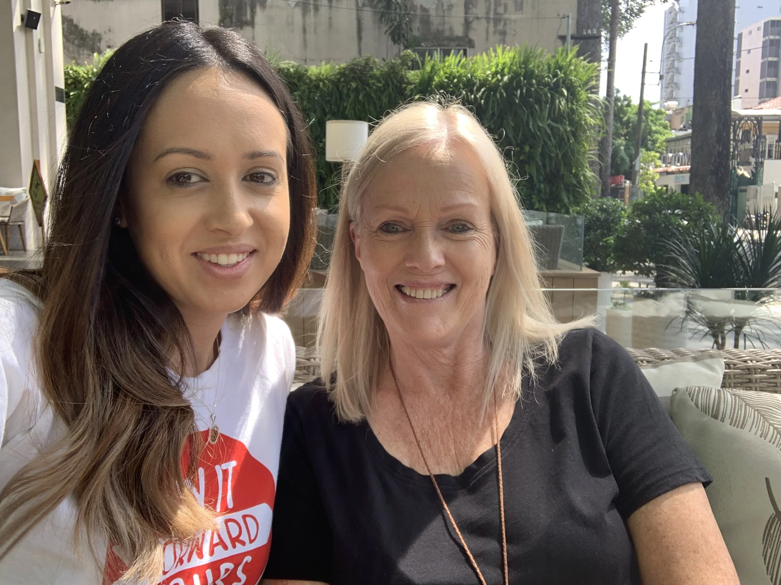 Two women smiling and sitting outdoors on a patio with greenery and buildings in the background.