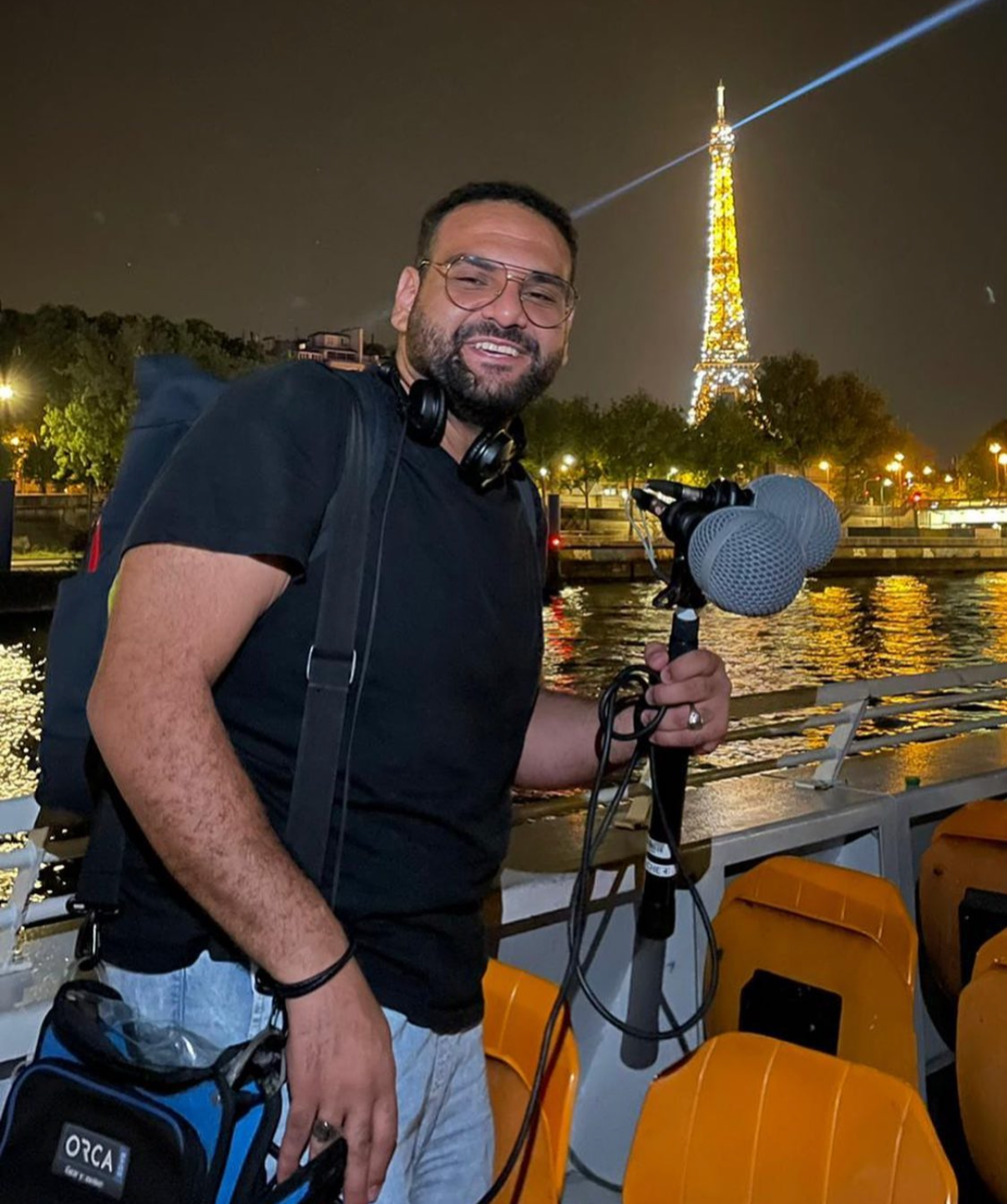 A man with a beard and glasses smiling at night on a boat near the Eiffel Tower, holding a microphone with foam covers.