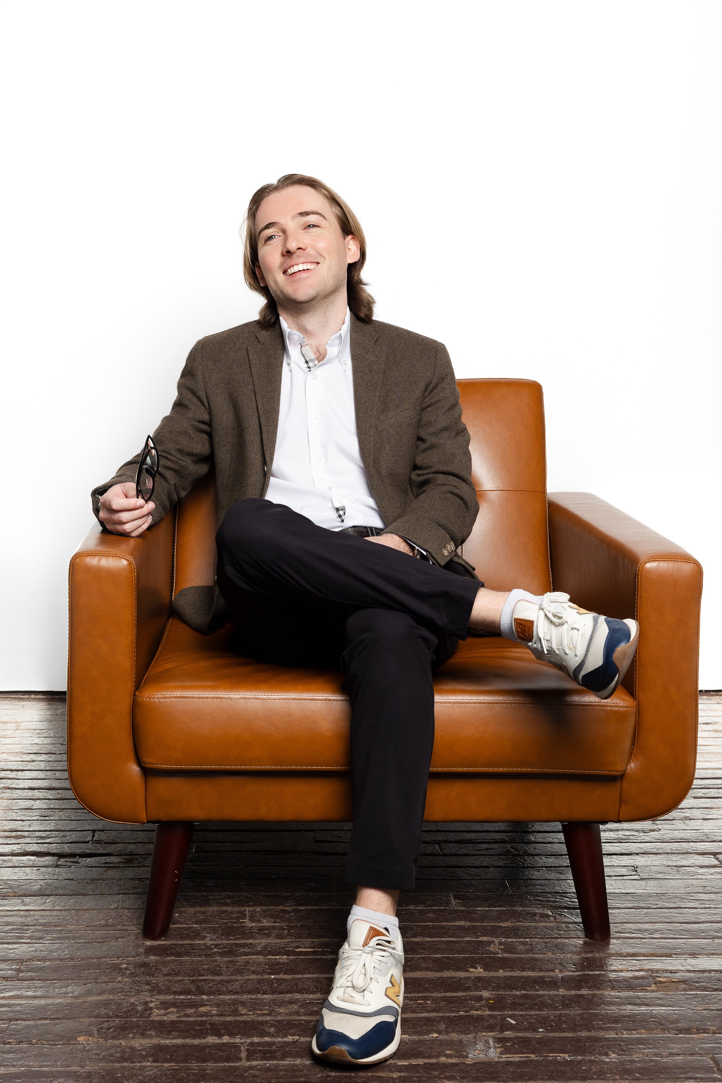 A man sitting on a tan leather armchair, smiling, holding glasses, in front of a white background.