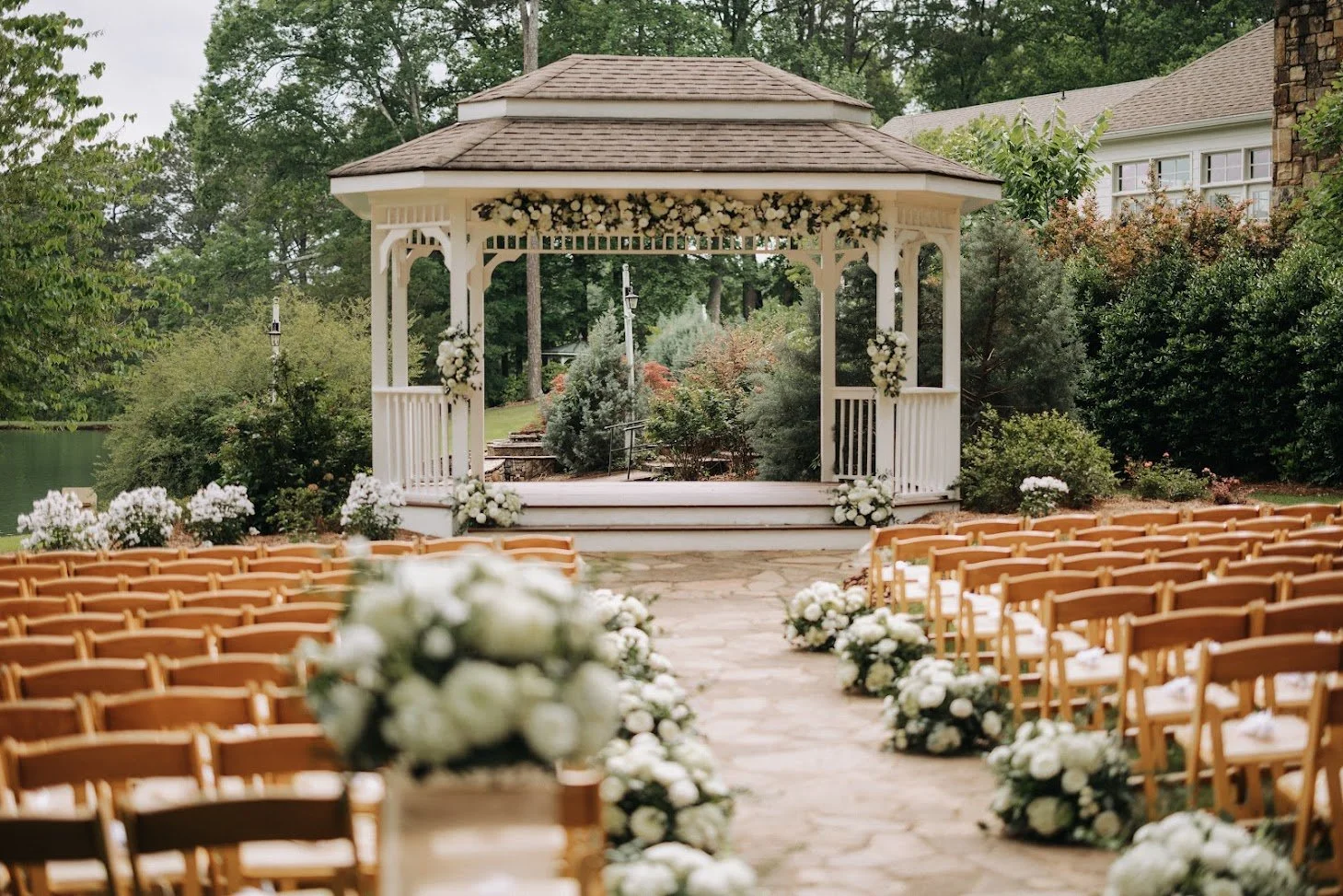 An outdoor wedding setup features rows of wooden chairs decorated with white flowers, facing a white gazebo with floral arrangements, in a lush garden setting near a pond.