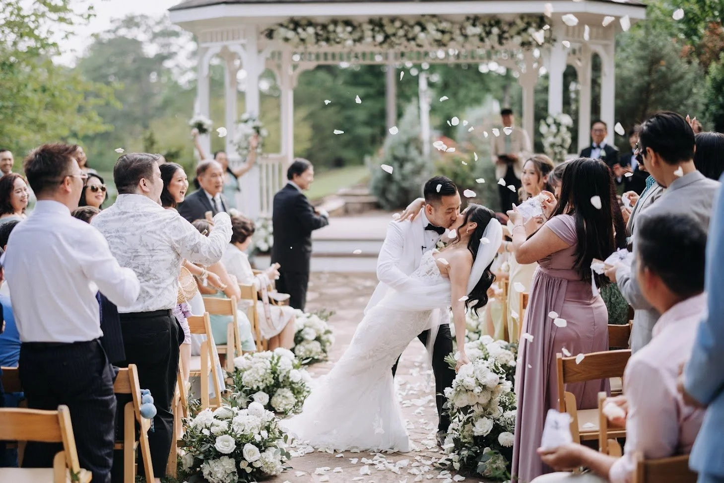 A wedding ceremony outdoors with a bride and groom kissing, surrounded by guests throwing flower petals under a white gazebo.