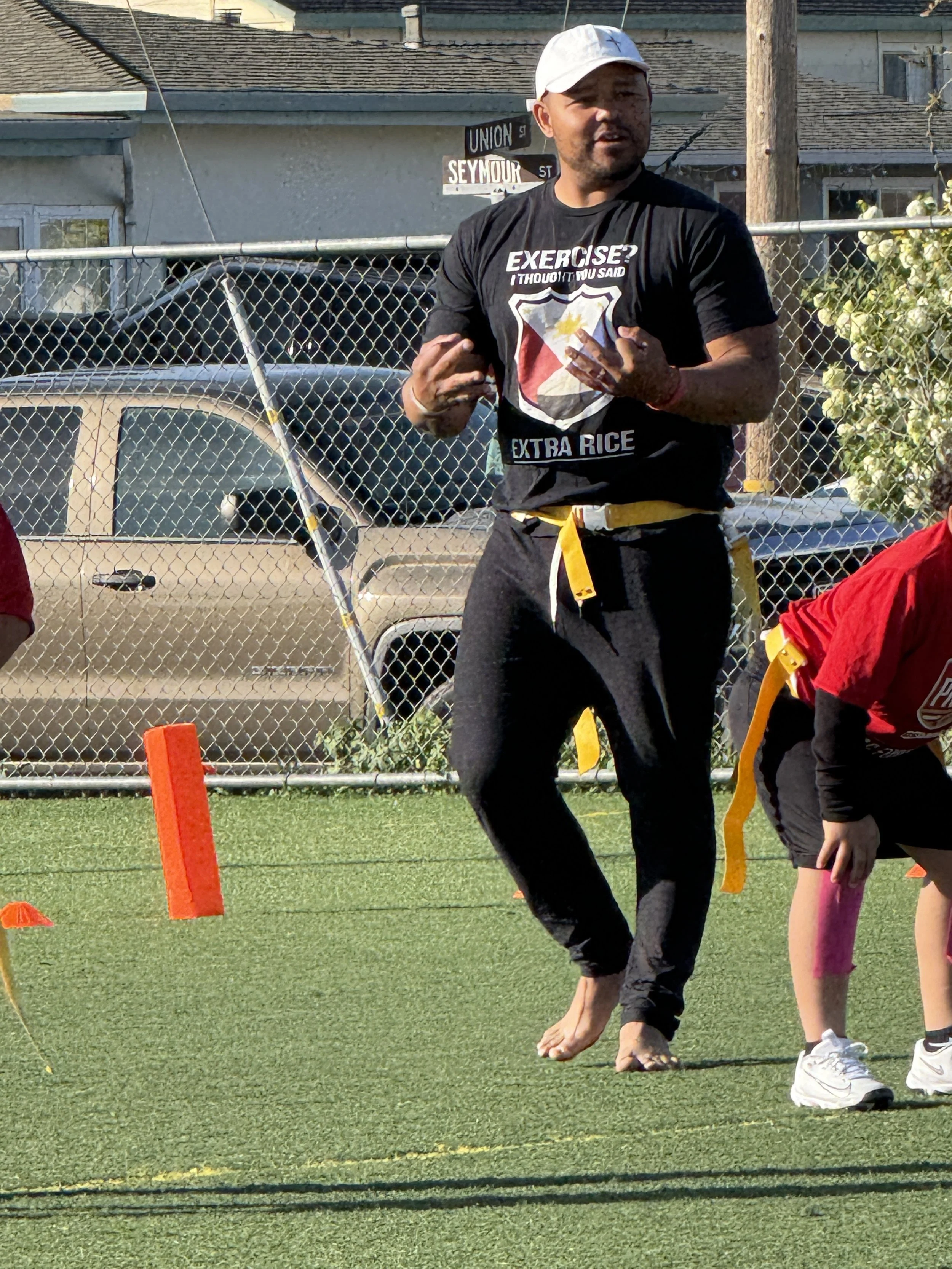 A man wearing a white cap, black T-shirt, and black pants barefoot, standing on a grassy field next to a child. The man is making a gesture with his hands, and the child is bending down. Orange cones are on the ground, and a chain-link fence with parked cars and house rooftops is in the background.