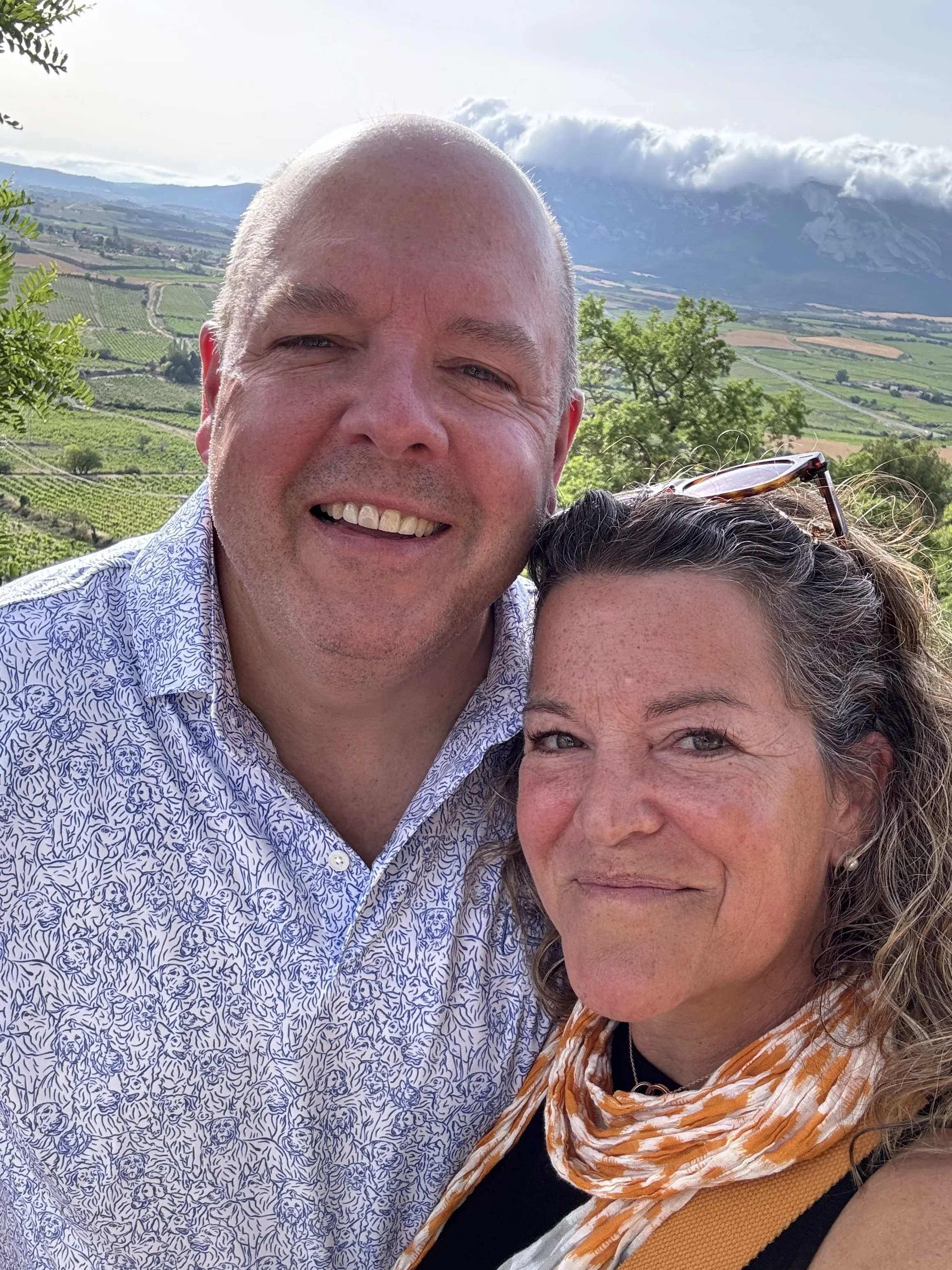 A smiling man and woman taking a selfie with a scenic landscape of vineyards, trees, and mountains in the background.