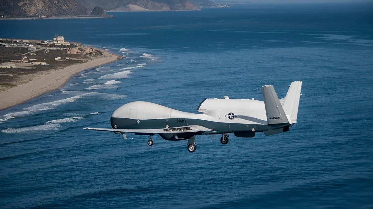 A military drone flying over the ocean with a shoreline and hills in the background.