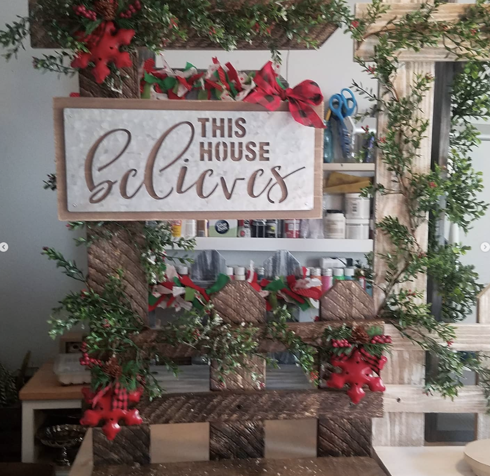 Decorative wooden display with a sign that reads 'This house celebrates'. The display is adorned with Christmas-themed decorations including red bows, green foliage, red poinsettias, pine cones, and red berries, alongside shelves holding various cont