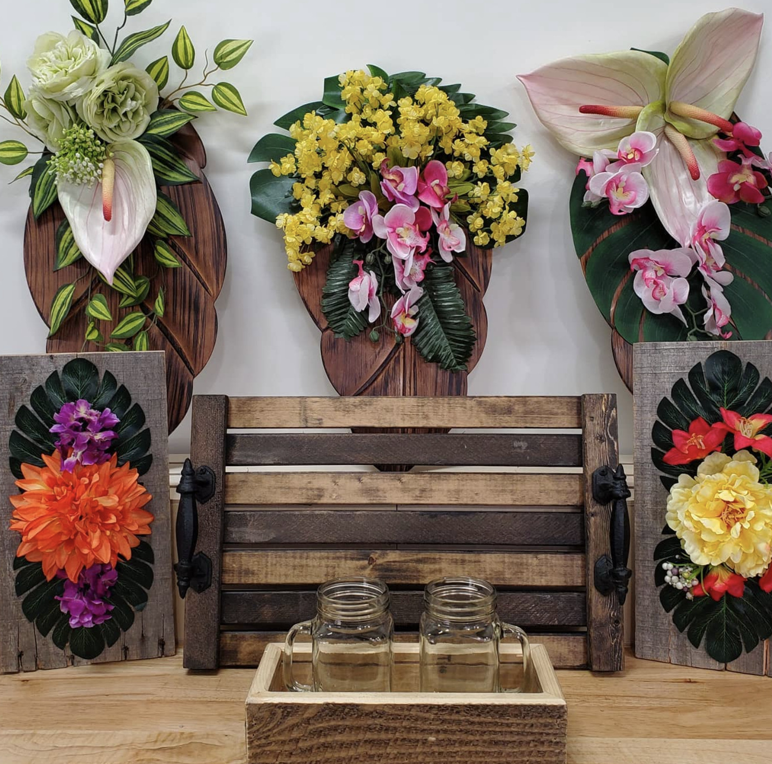 Decorative floral wall arrangements with wooden backgrounds on a tabletop, including yellow, pink, purple, orange, red, and white flowers, alongside two glass jars and a wooden tray.