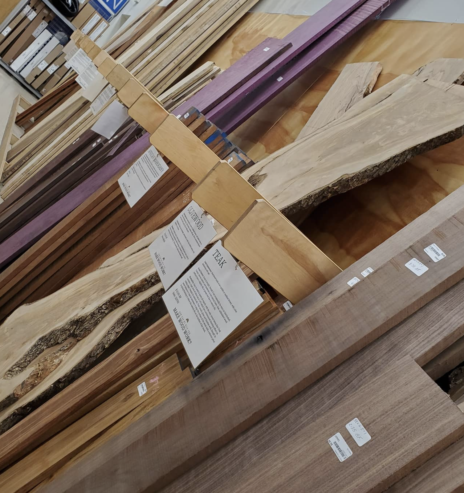 Various types of wood planks and boards on display at a store, including natural, stained, and painted wood pieces with informational tags.
