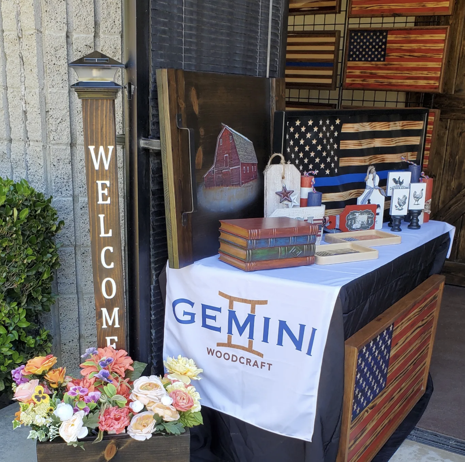 Display table with patriotic-themed decor, signs, and crafts, featuring American flags, a flower arrangement, and woodworking items, with a wooden 'Welcome' sign.