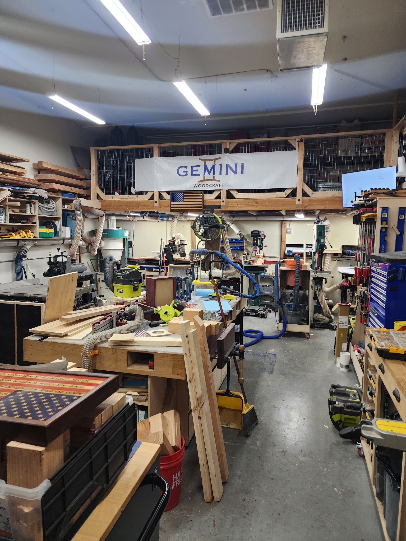 A woodworking workshop filled with tools, materials, and machinery, with a loft area above featuring a sign that reads 'GEMINI Woodcraft' and an American flag.