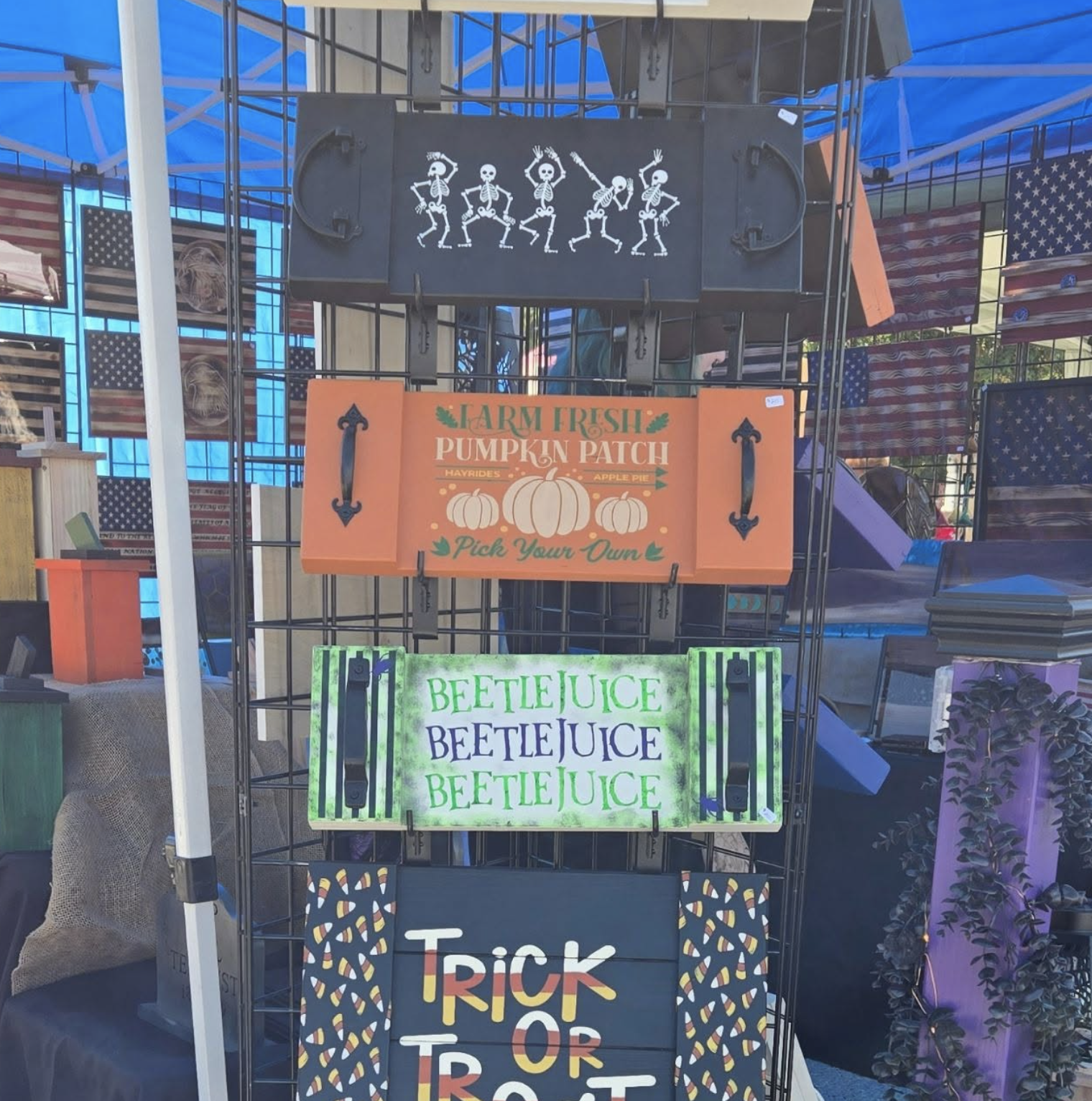 Display stand with Halloween and fall-themed signs including a sign for a pumpkin patch, a neon sign with dancing skeletons, and a sign for beet juice, at an outdoor market.