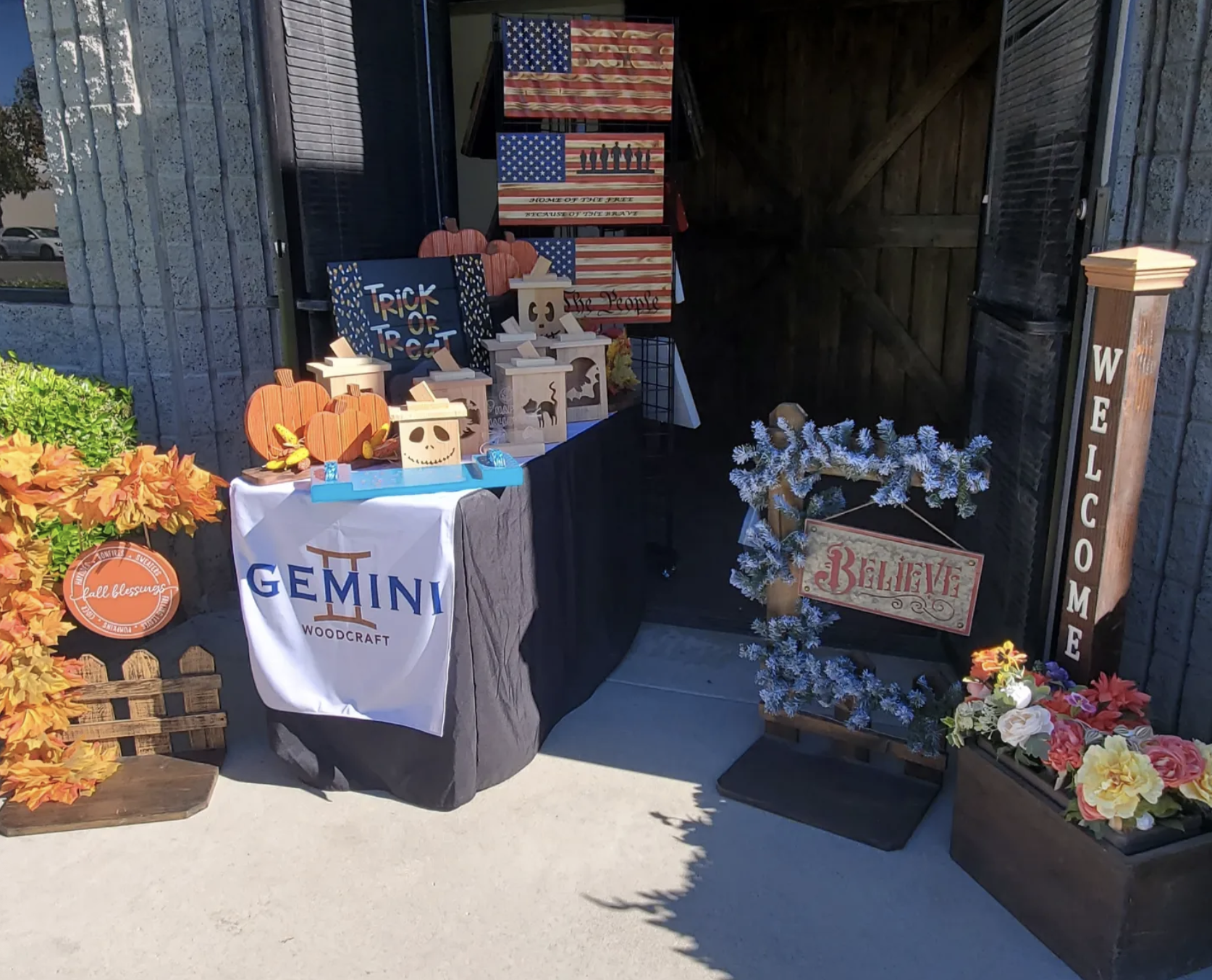 A patriotic display booth with American flags, fall decorations, and woodcraft signage, featuring containers with pumpkins, autumn leaves, and a welcome sign.