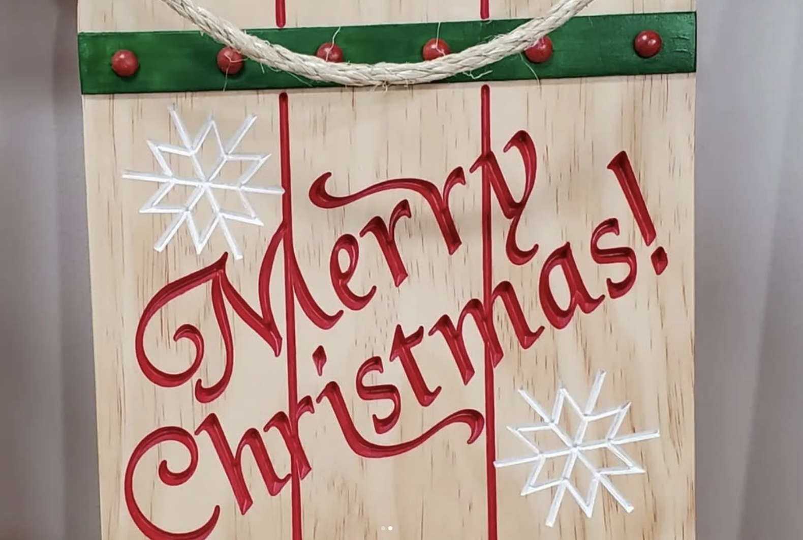 A decorative holiday sign with a wooden background, displaying the message 'Merry Christmas!' in red cursive letters. The sign is embellished with white snowflake lights, red ornamental accents, and a festive green and red rope at the top.