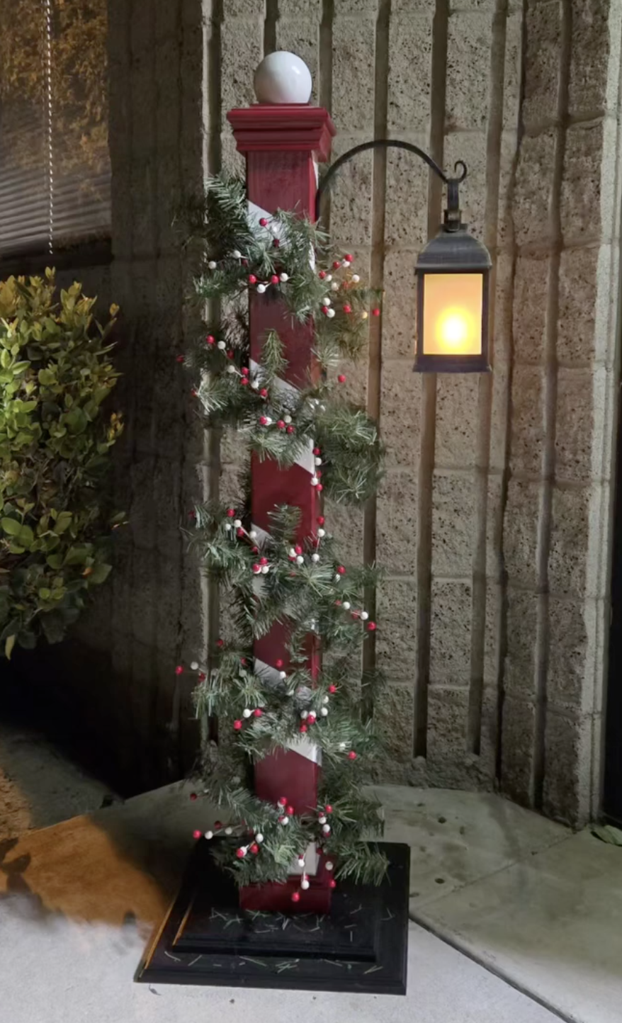 Decorated porch post with garland, red and white ornaments, and a lit outdoor lantern against a brick wall.