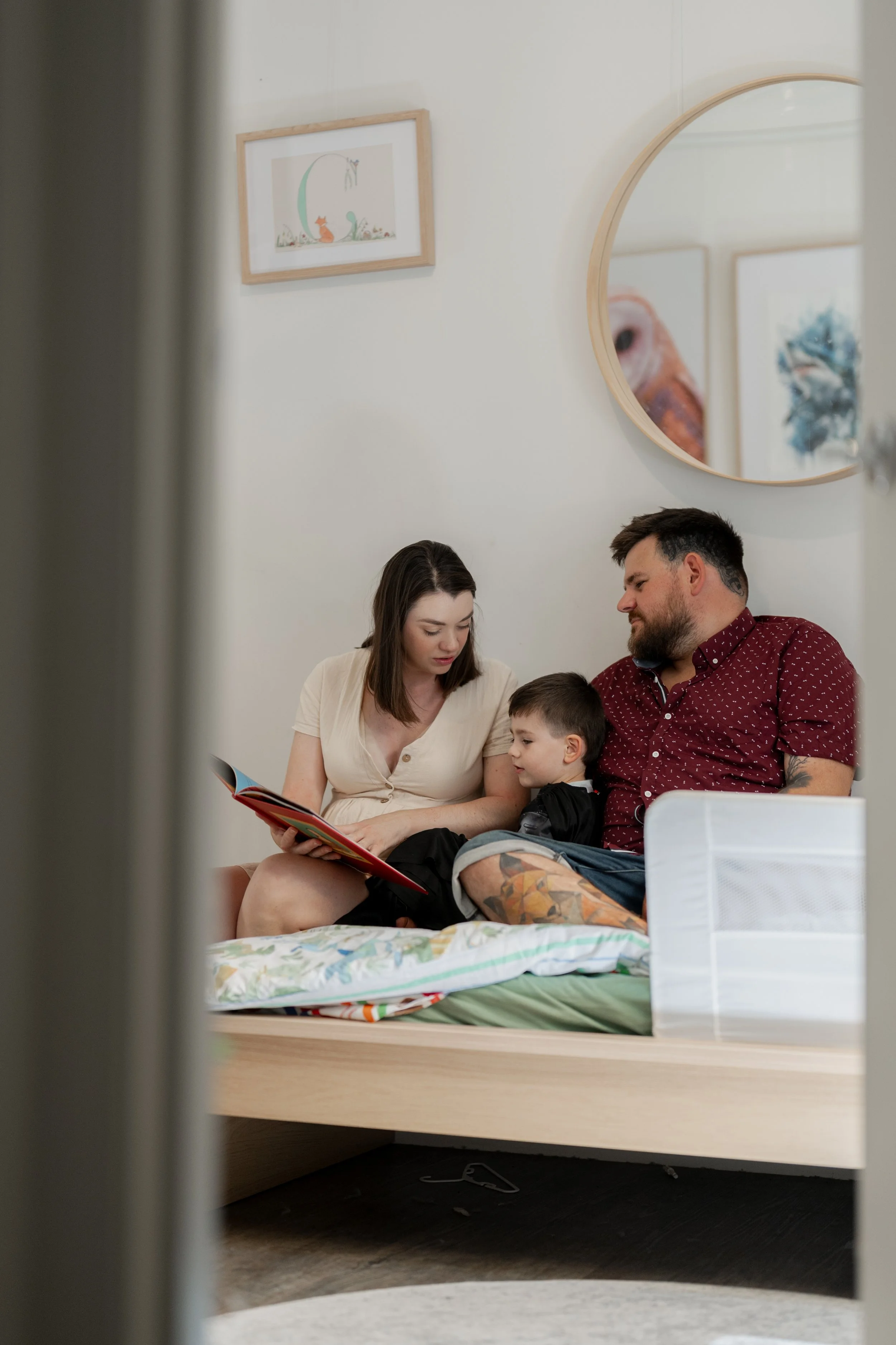 A family of three sitting on a bed, reading a book together in a cozy bedroom with framed pictures and a round mirror on the wall.