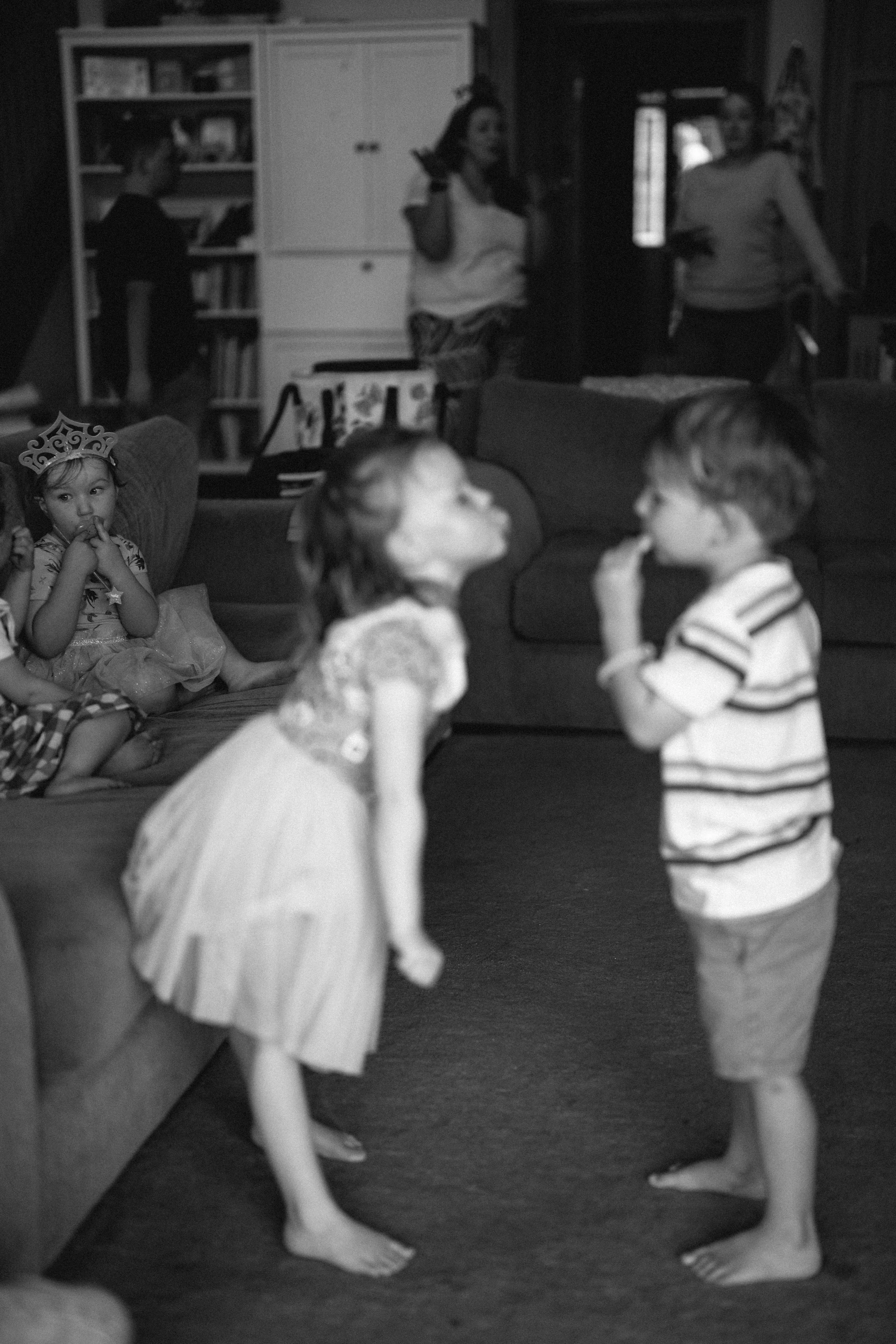 Children at a birthday party, with two kids standing and talking in the foreground, one girl wearing a princess crown and a girl seated on the couch wearing a princess costume, in a room with bookshelves and adults in the background.