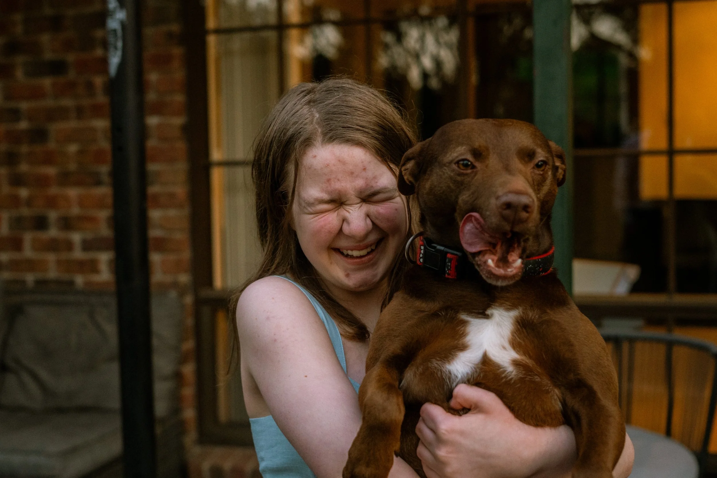 A girl with glasses hugs a brown dog with a wide, open-mouthed smile. They are outside near a brick building.