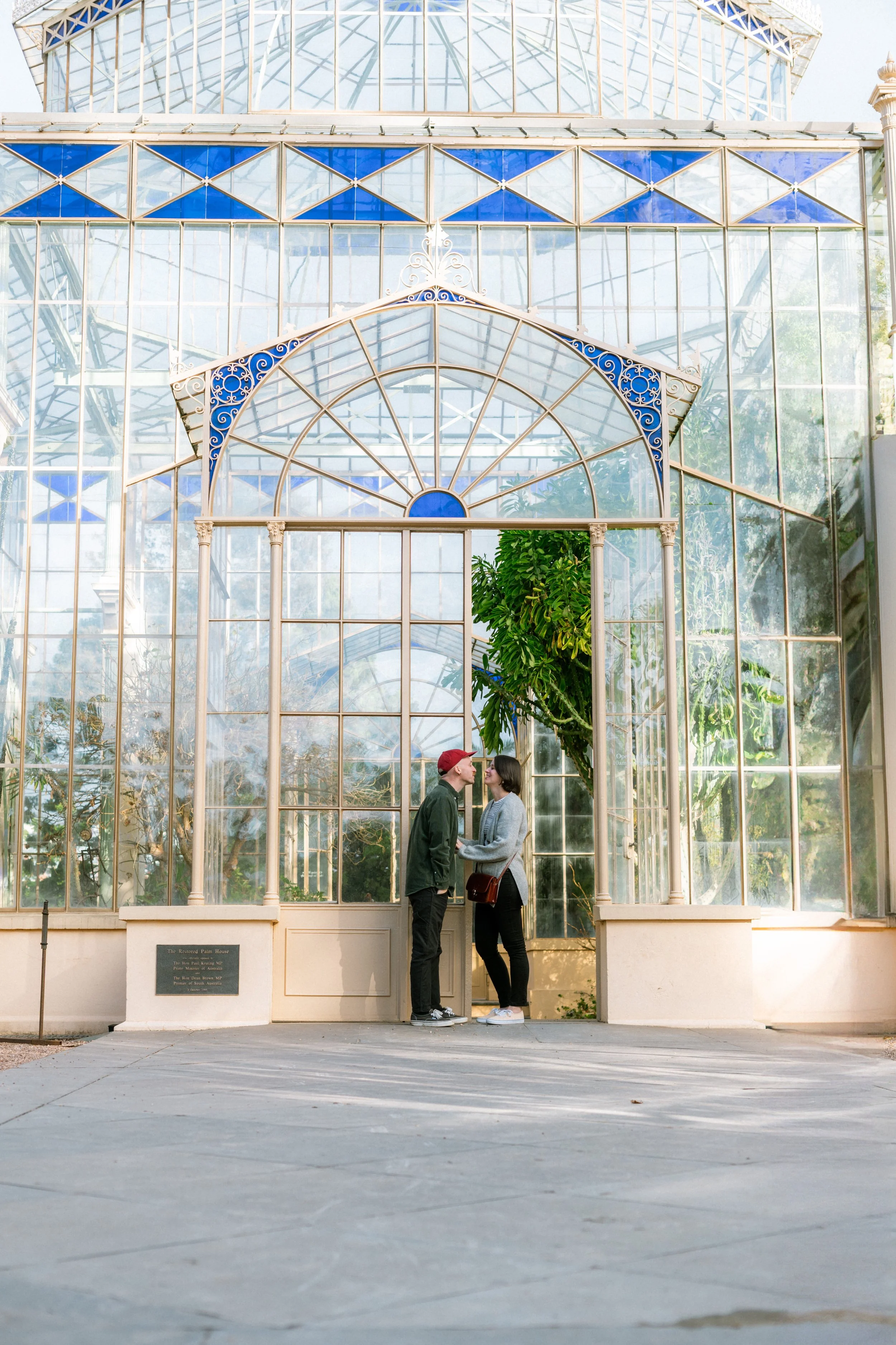 A couple stands close to each other in front of a large glass greenhouse, with ivy in the background and a plaque on the wall to the left.