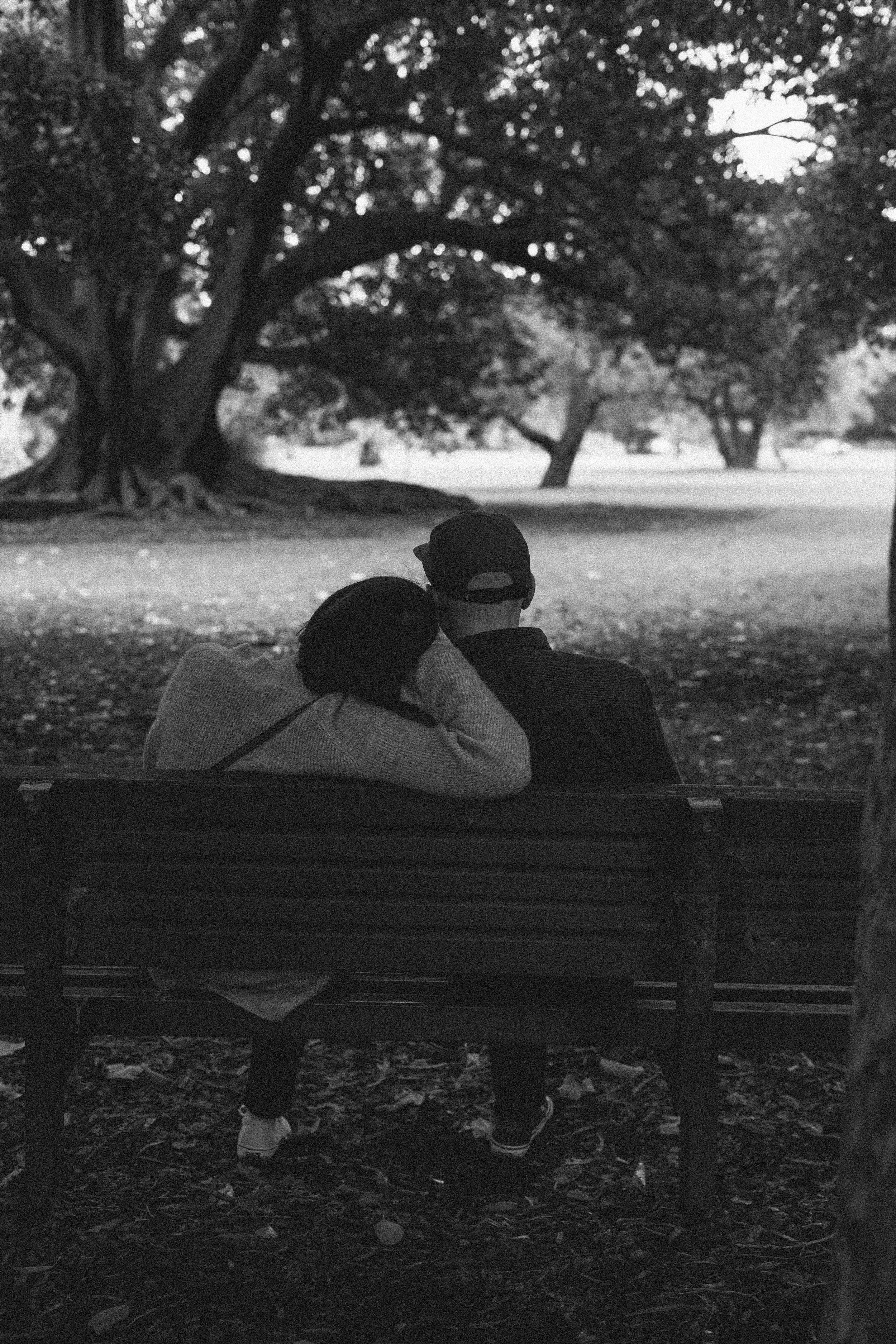 A black and white photo of a couple sitting on a park bench, with the woman resting her head on the man's shoulder. They are seated under large trees in a peaceful park setting.