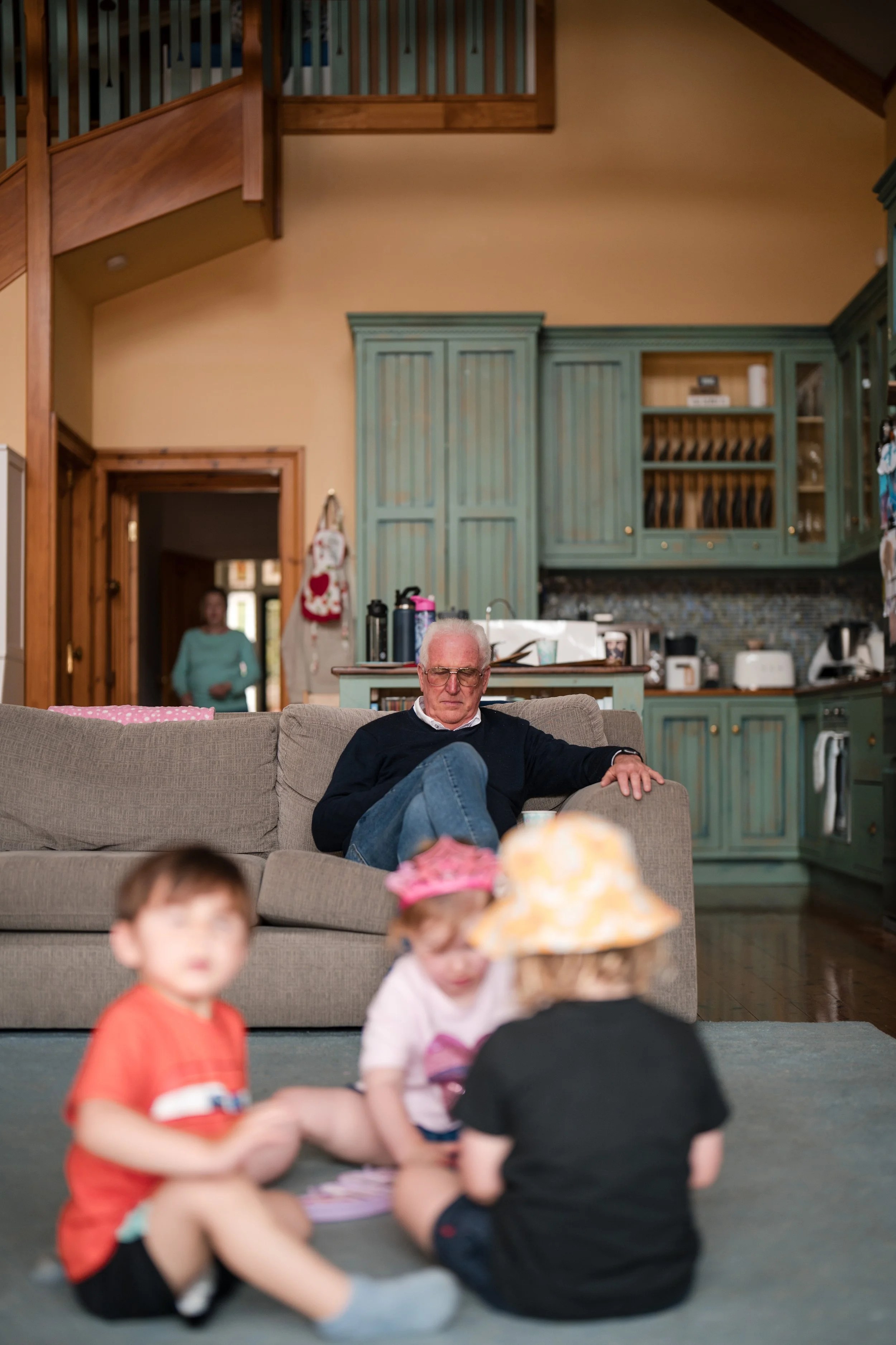 An elderly man sits on a couch watching children playing on the floor in a kitchen and living room area.