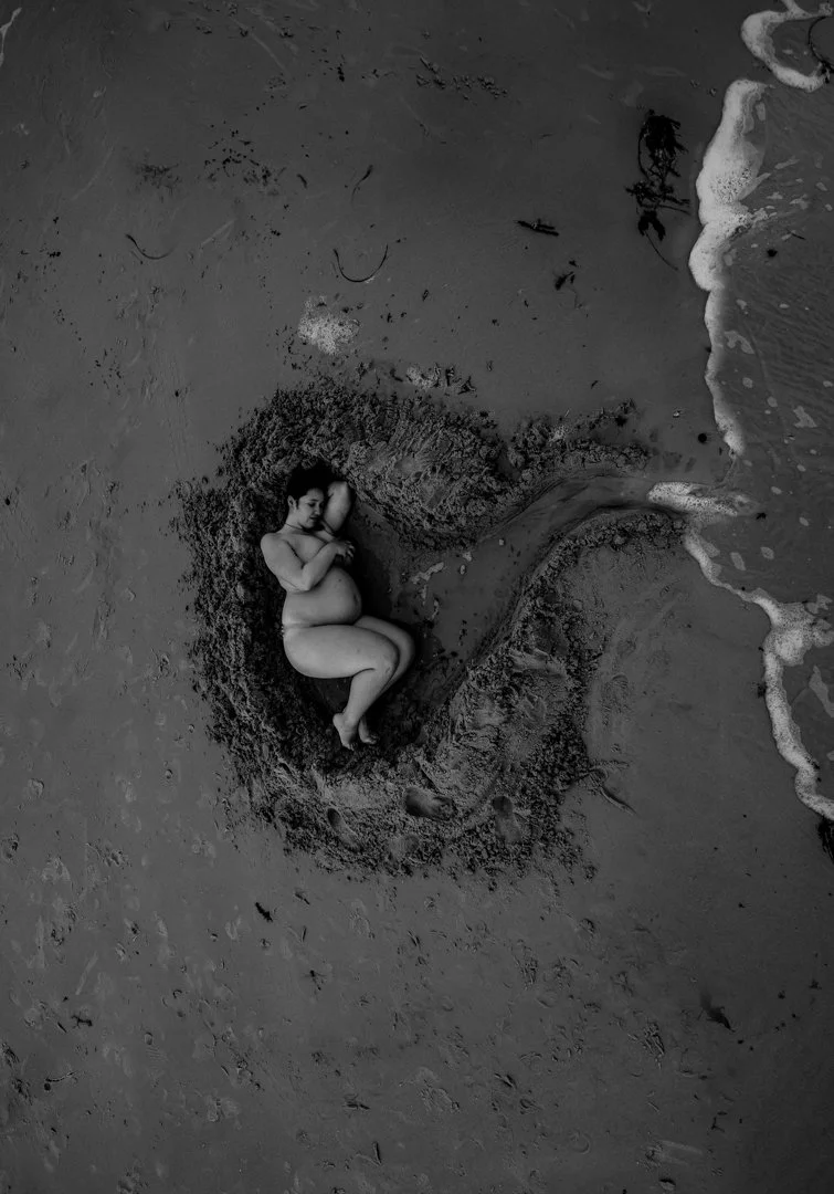 A black and white photo of a pregnant woman lying in a heart-shaped sand cavity on a beach near the water's edge.