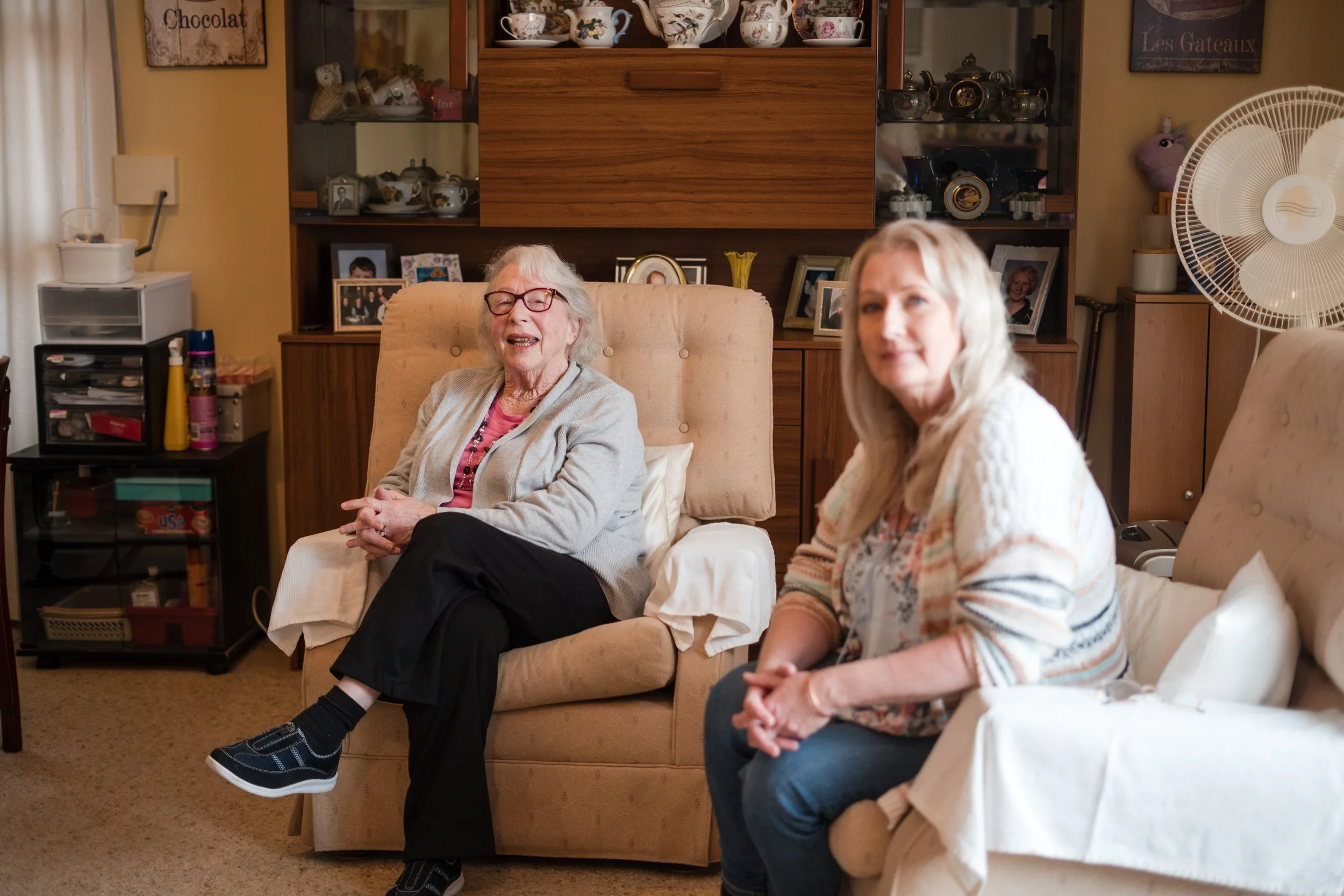 Two women sitting on couches in a cozy living room, smiling and engaging in conversation, with a wooden cabinet and personal photos in the background.