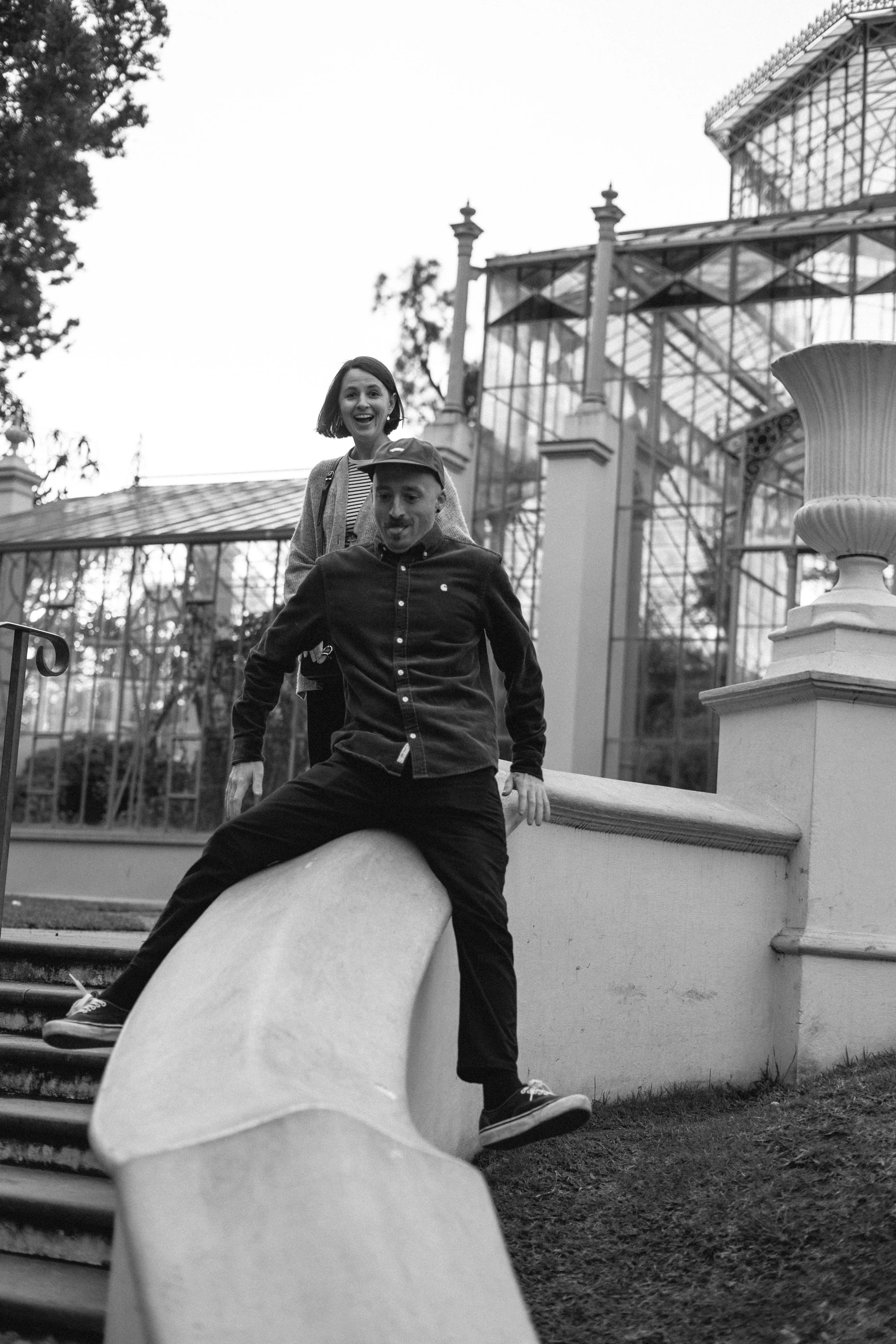 Black and white photo of two people, a woman and a man, on a slide outside a glass greenhouse. The woman is smiling in the background, and the man is sitting on the slide in the foreground.