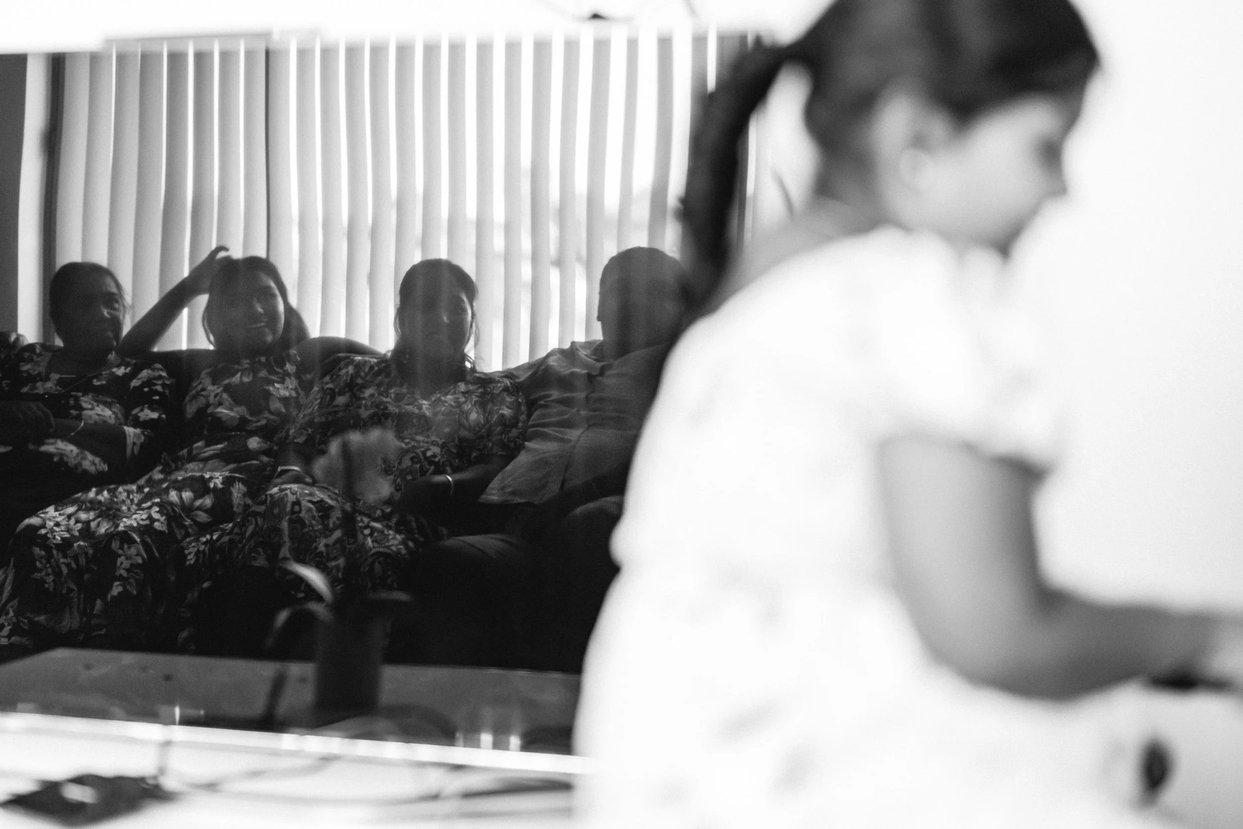 A woman, seen in profile, is sitting at a table with her arms resting on it. In the background, a group of people is seated on a sofa, smiling and engaging with each other, with vertical blinds covering the window behind them.