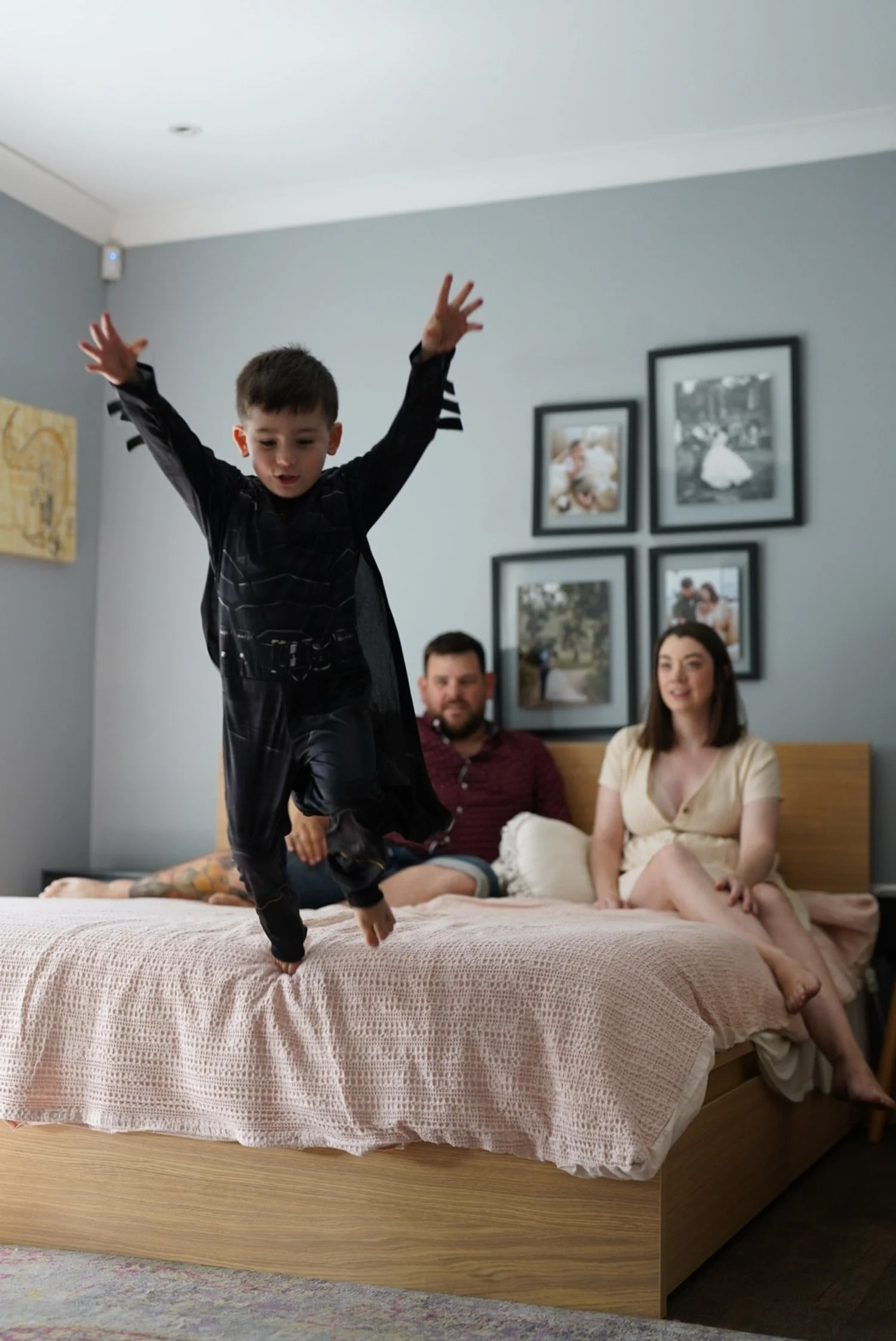 A young boy in a black superhero costume is jumping onto a bed with a pink blanket, while a woman in a yellow dress and a man with a beard sit on the bed watching, in a bedroom with gray walls and framed photos.