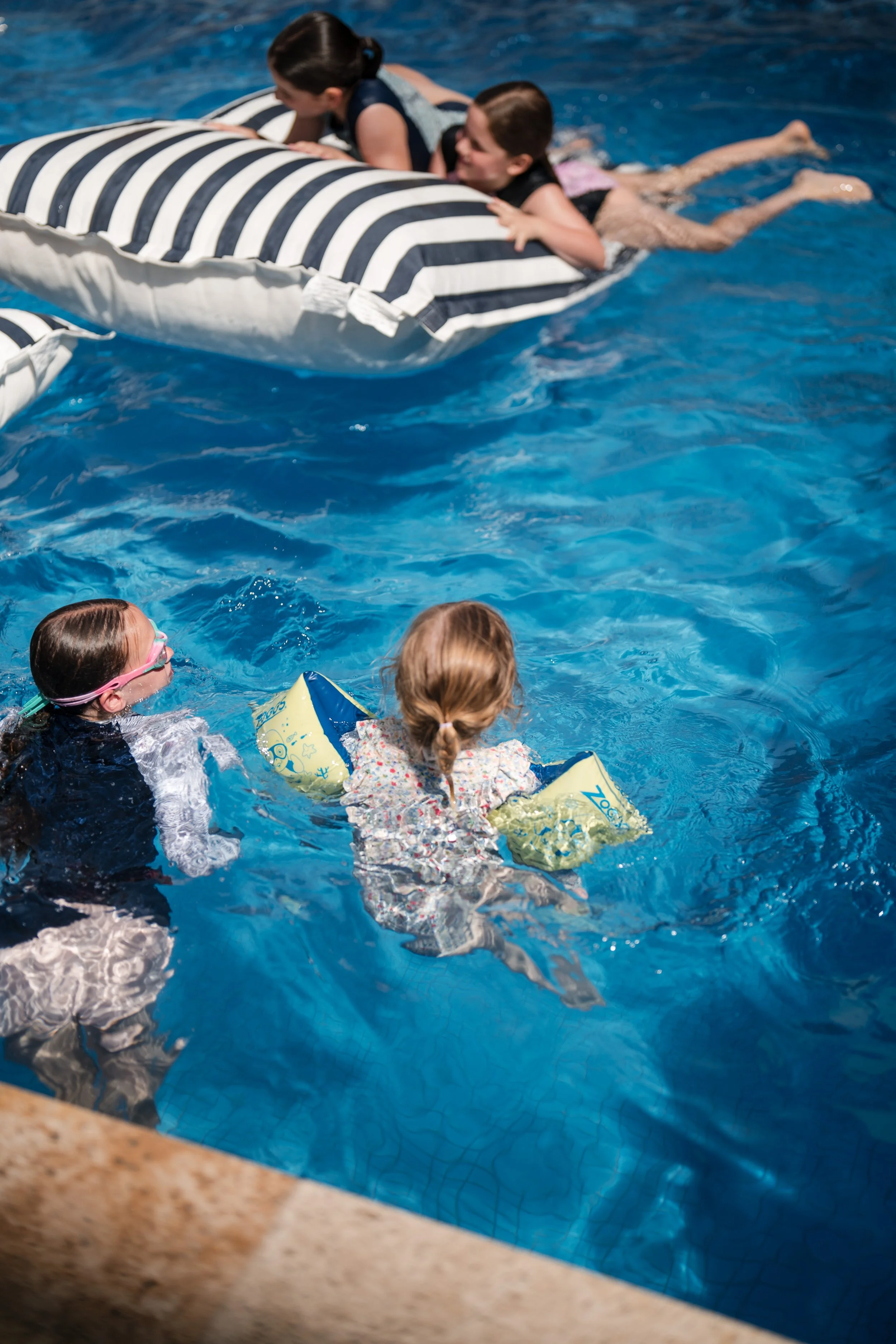 Children swimming in a pool, with some children on a striped inflatable float and others using floatation devices.
