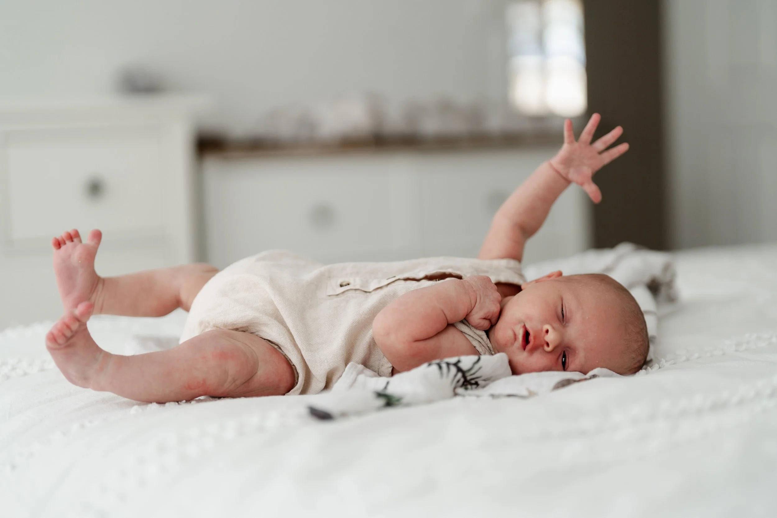 A newborn baby lying on their side on a white bed, reaching with one arm raised, in a cozy room with white furniture and a window in the background.