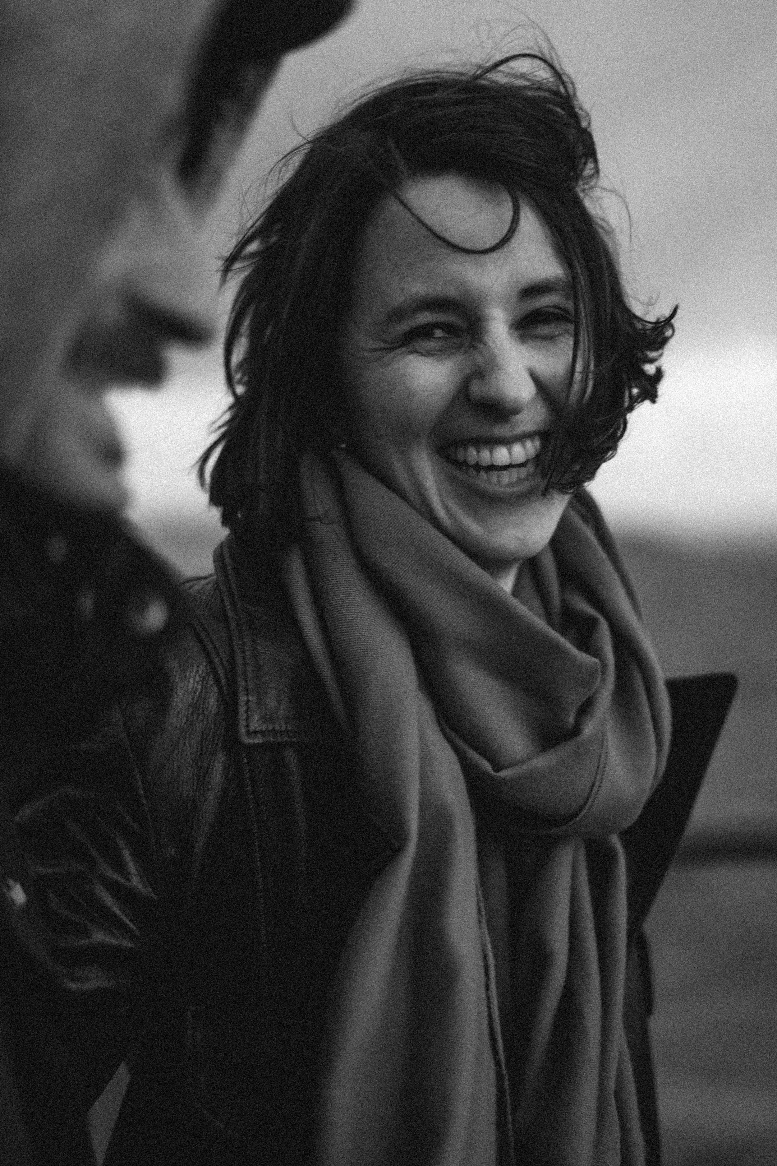 A woman smiling and laughing, wearing a scarf and leather jacket, outdoors in black and white.