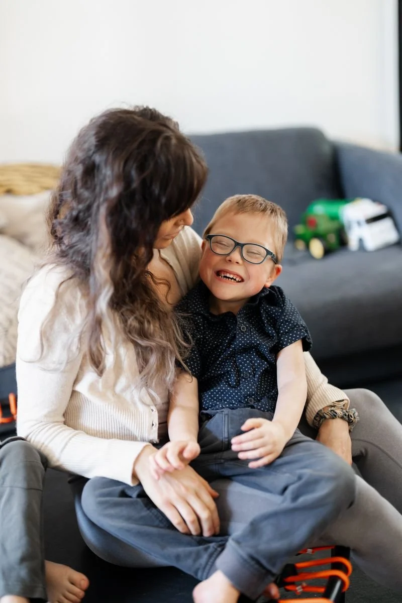 A woman and a young boy sharing a joyful moment, with the boy wearing glasses and smiling widely, sitting on a couch in a living room.
