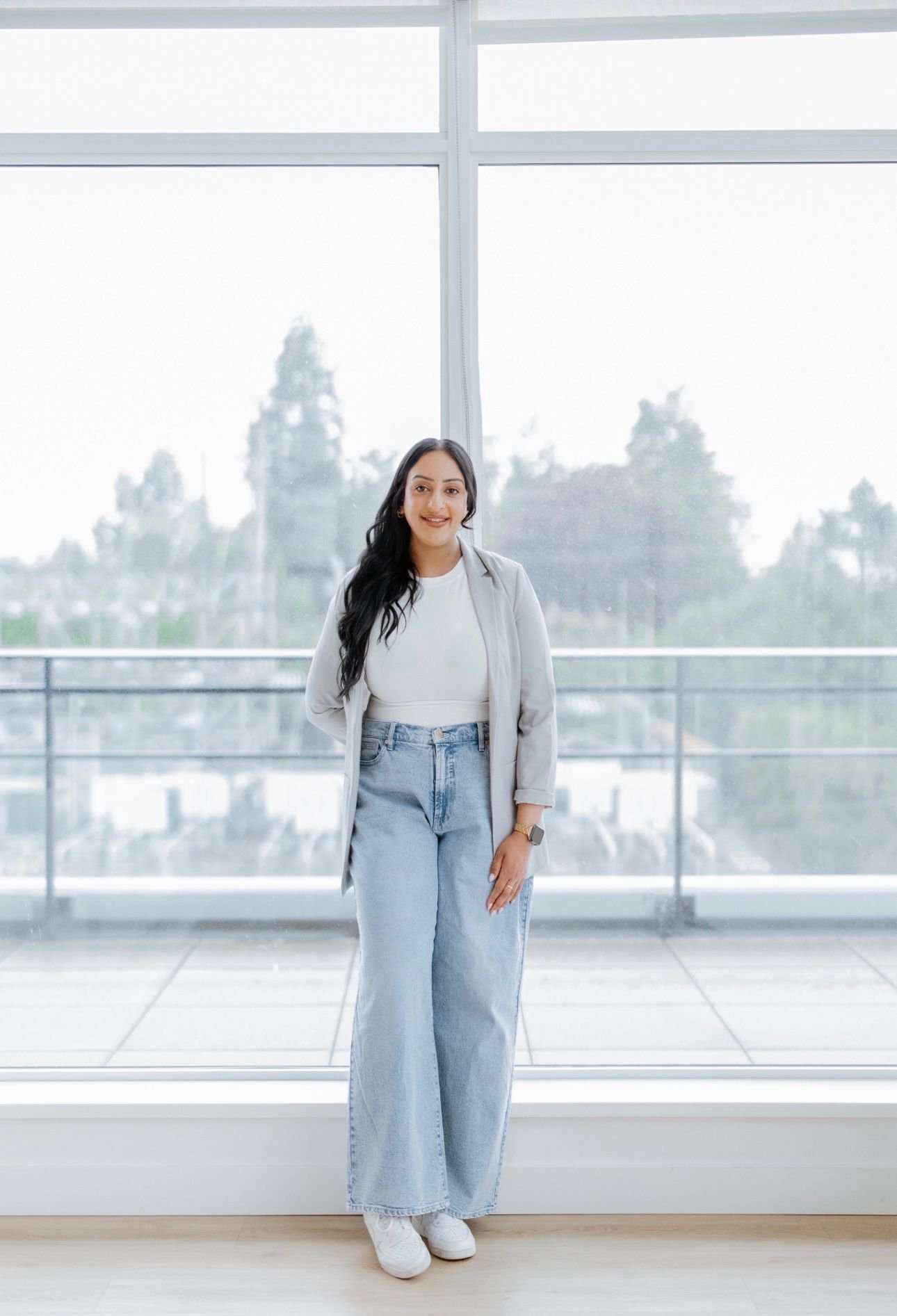 A woman with long brown hair and jewelry, smiling, sitting at a table with a white tablecloth, decorated with plates, utensils, a napkin, and a glass of champagne, in a warmly lit setting.