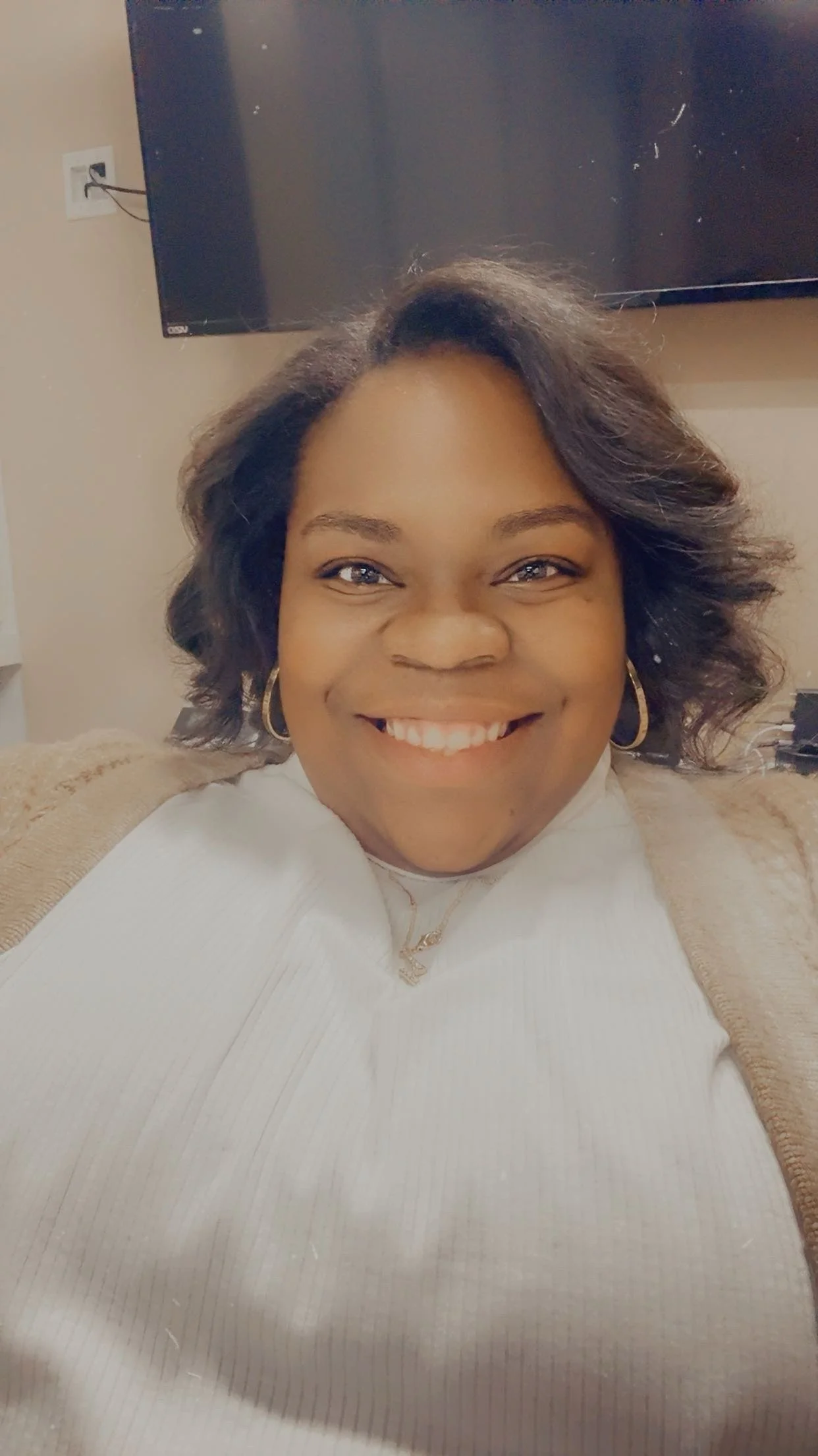 A smiling woman with shoulder-length dark hair, wearing hoop earrings and a white top, sitting on a beige couch in front of a television.