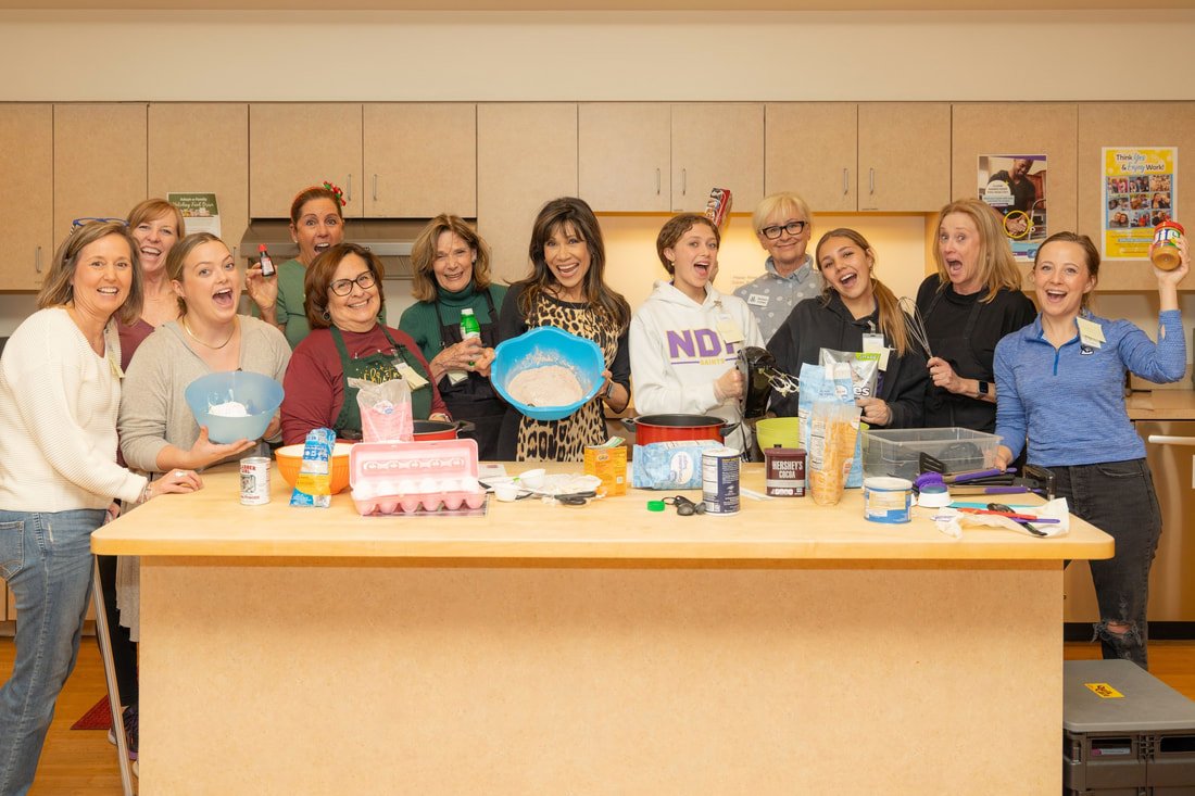 Group of twelve women smiling behind a kitchen counter with baking ingredients and tools, celebrating or preparing for a baking activity.
