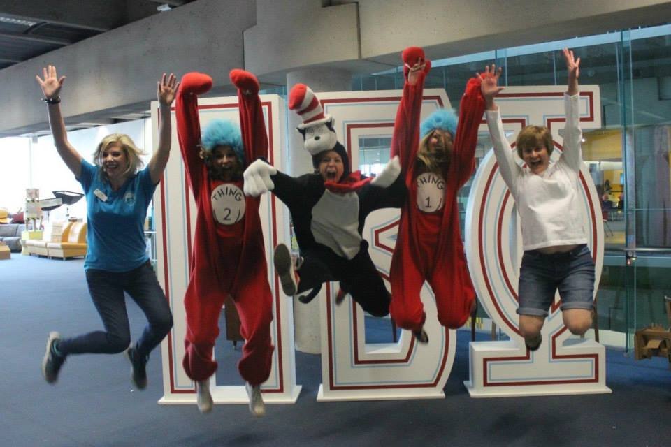 Four people are jumping in front of large 'MO' letters, with two individuals dressed as Thing 1 and Thing 2 from Dr. Seuss, and the other two women smiling and raising their hands, inside a modern building.