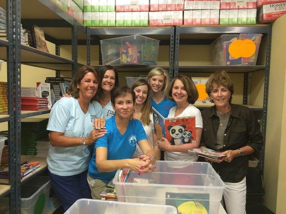 Seven women smiling, standing and sitting in a room with metal shelving filled with books, binders, and plastic containers, posing for a group photo.