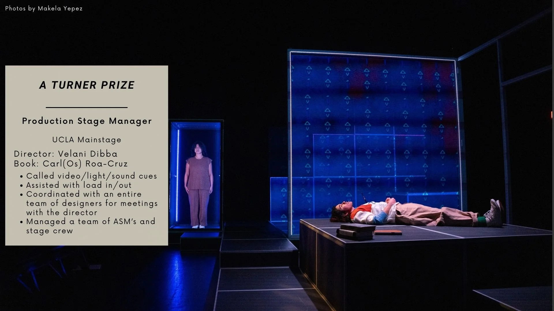 The image shows a theatrical stage setup with a female actor lying on the stage and another woman standing in a blue-lit box frame. There is a large translucent screen with geometric patterns in blue lights behind them. On the left side of the image,