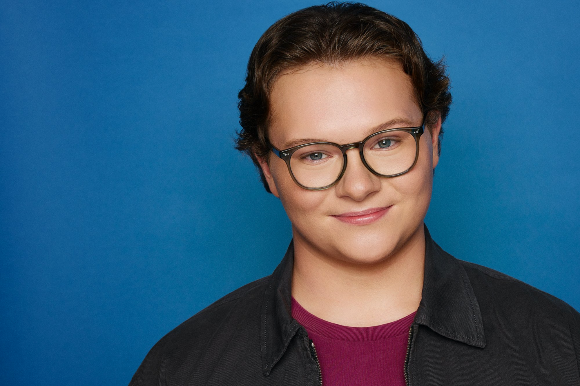 A young person with short brunette hair, wearing glasses, a black jacket, and a maroon shirt, smiling slightly against a blue background.