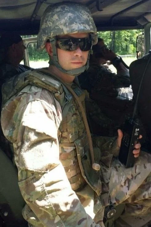A soldier in camouflage uniform and helmet sitting in a vehicle, holding a radio, wearing sunglasses.