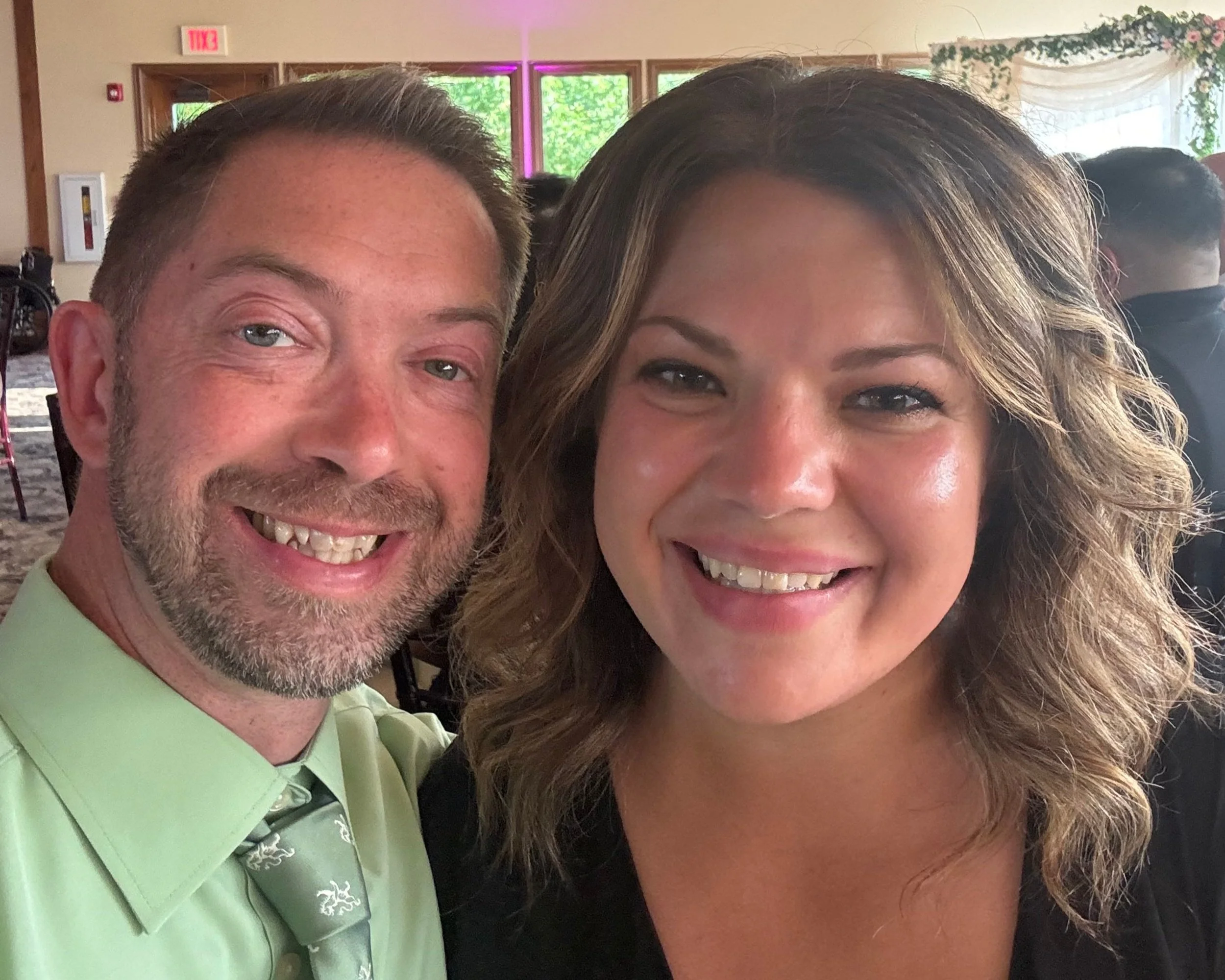 A smiling man and woman taking a selfie at an indoor event. The man has short hair and a beard, wearing a light green shirt and patterned tie. The woman has shoulder-length wavy hair and is wearing makeup and a black top.