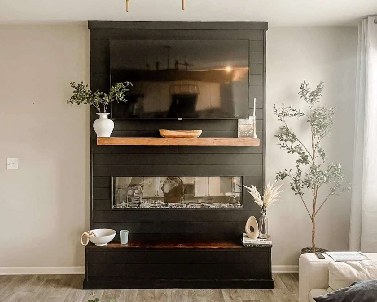 Living room wall with a mounted flat-screen TV above a black shiplap panel and a rectangular glass fireplace. A wooden mantel holds a white vase with greenery, a wooden bowl, and a candle. Below the mantel, there are decorative items, including a bowl, a mug, and a small sculpture, on a dark wood cabinet. To the right, a tall houseplant stands next to a white sofa.