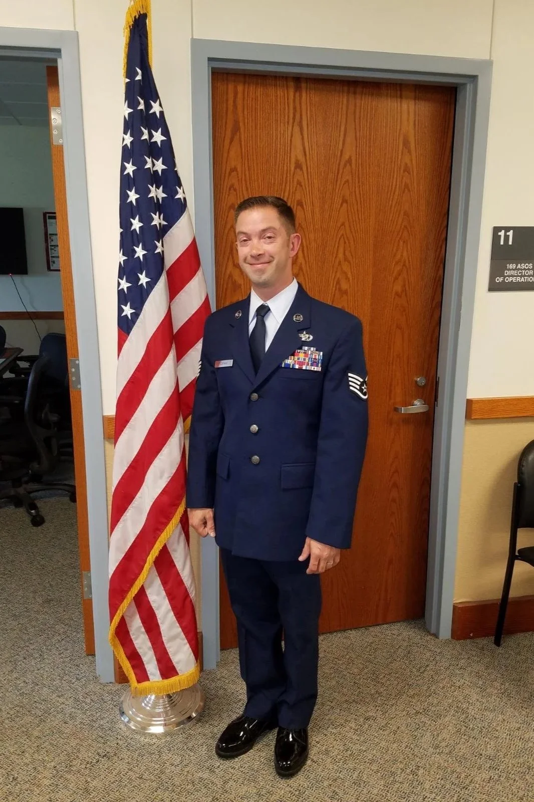 A man in a U.S. Air Force dress uniform standing next to an American flag in an office setting.