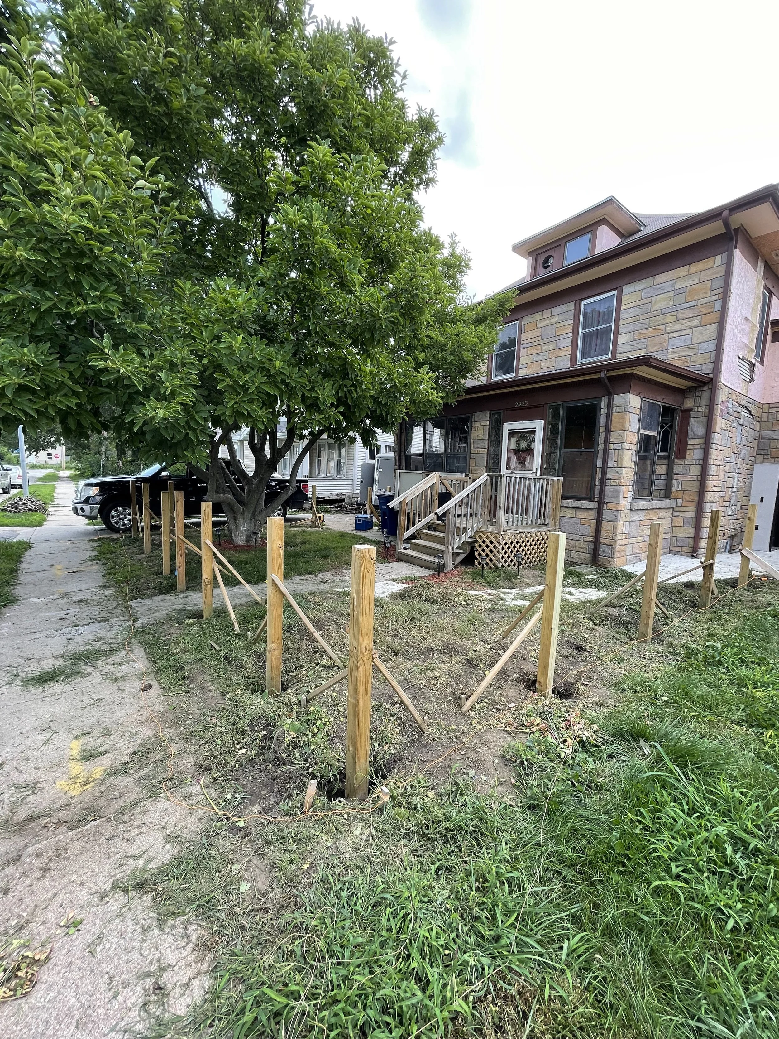 A house with construction in progress shows new wooden fence posts installed around a yard, with a large green tree and parked cars nearby.