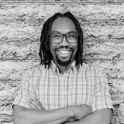 DeMonte Jackson, owner of Enhance Remodeling, smiling confidently with arms crossed in front of a textured wall, wearing glasses and a checkered shirt.