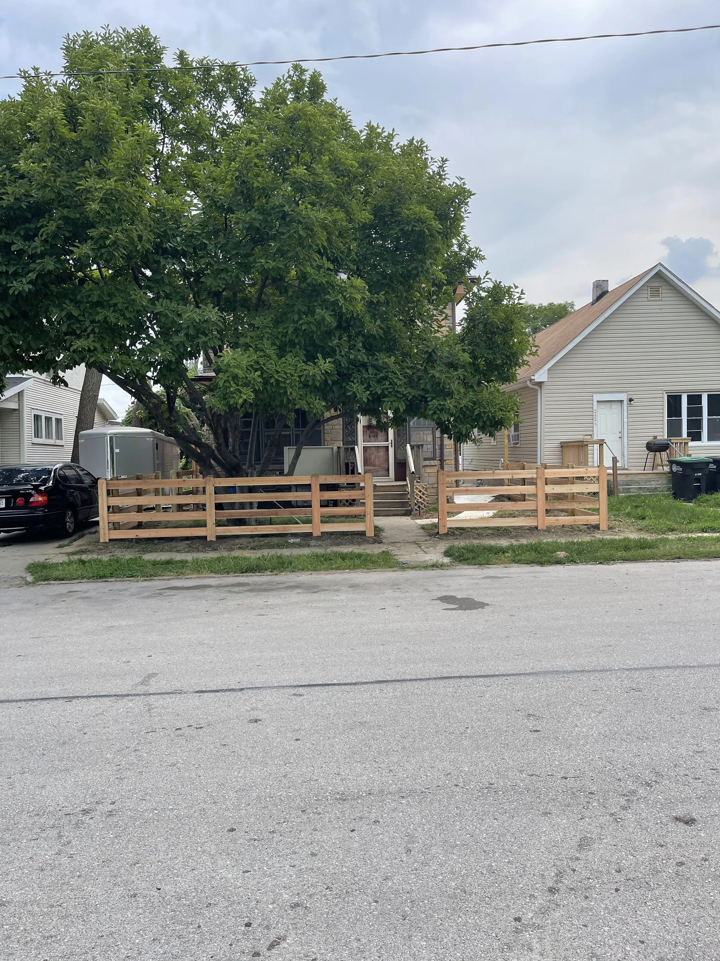 A house with a large tree in front, a wooden fence, a black car, and a trailer on the driveway. The house has beige siding, a front porch, and a small deck with stairs.