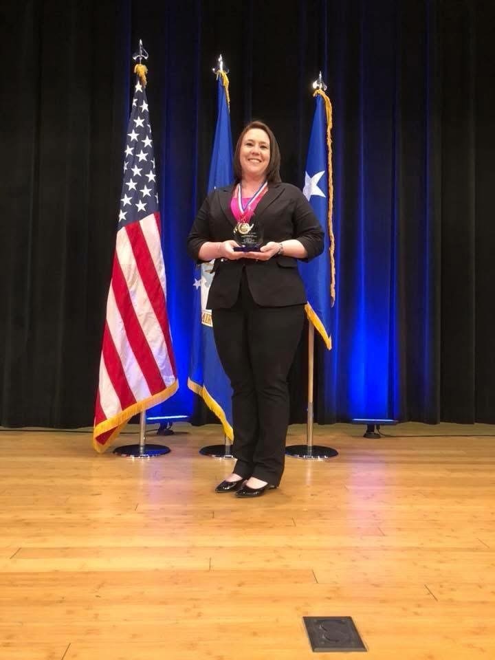 A woman in a black suit standing on a stage, holding an award medal, with American and Texas flags behind her.