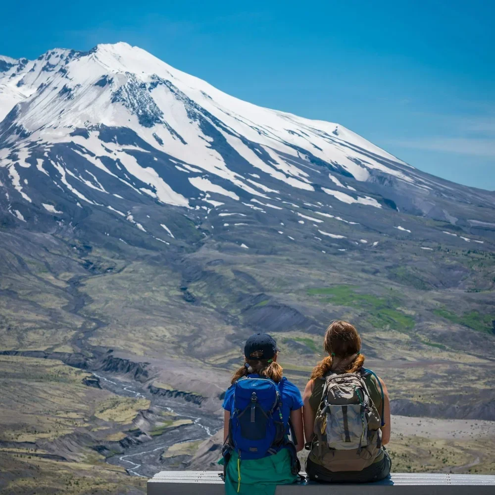 Two people with backpacks sitting on a bench, looking at a snow-capped mountain.