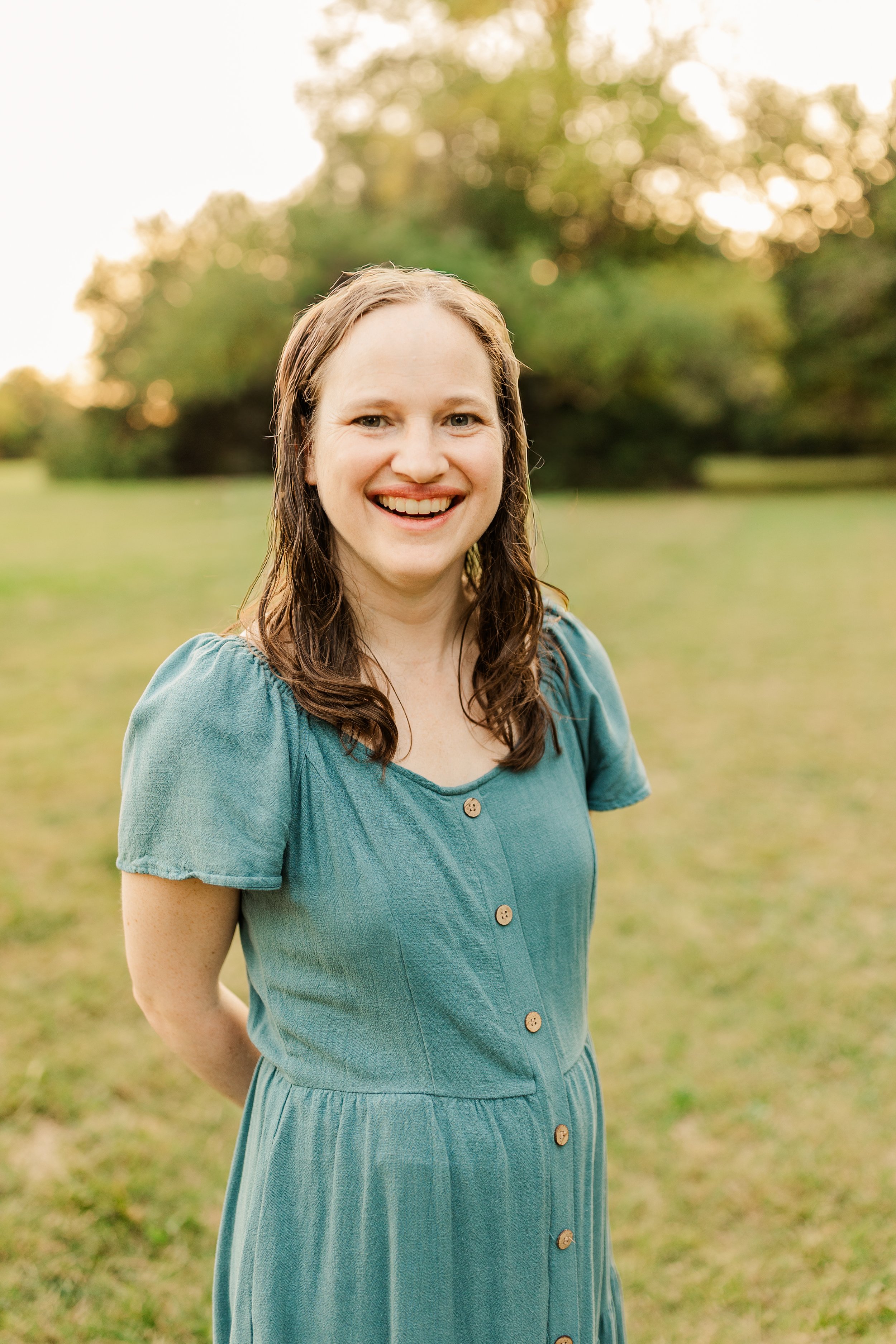 A smiling woman with shoulder-length brown hair standing outdoors in a grassy field with trees in the background during sunset.