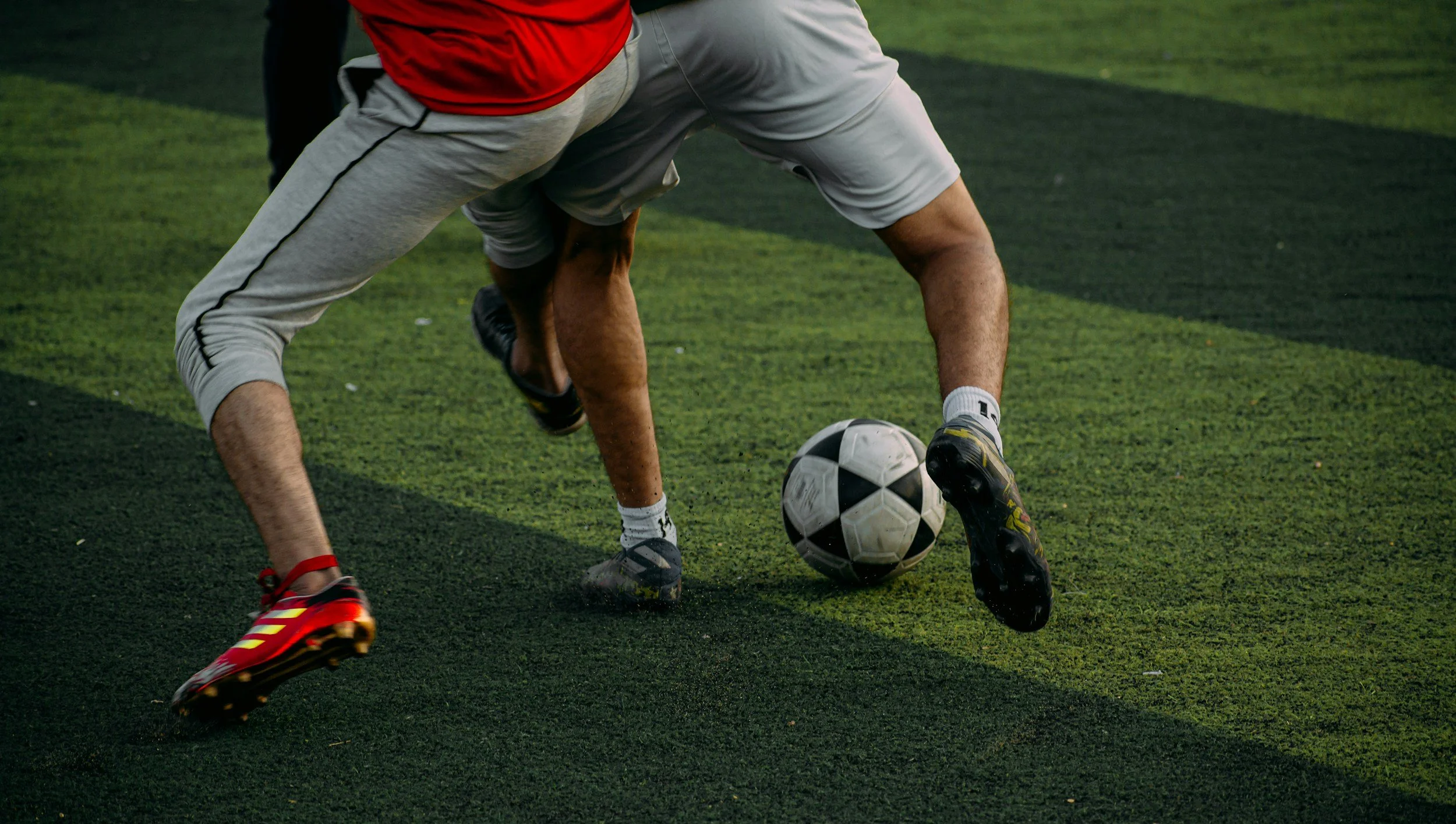 A person playing soccer, wearing a red shirt, gray shorts, and cleats, is in a crouched position on a green artificial turf field, with a black and white soccer ball nearby.